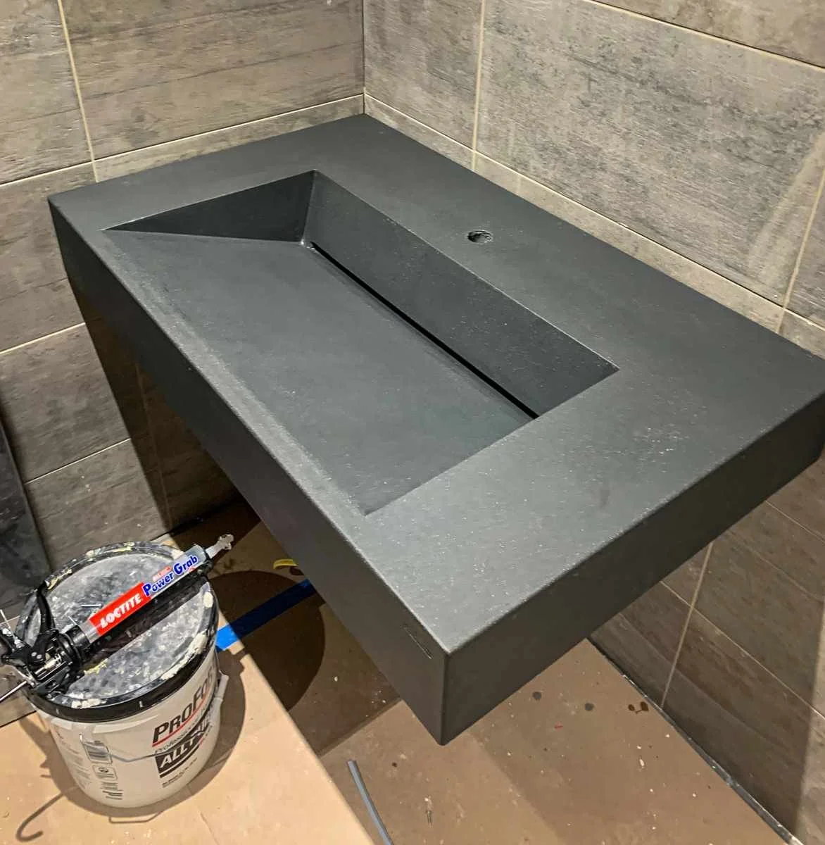 A black rectangular sink mounted on a wall with beige tile. There is a bucket with tools below the sink.