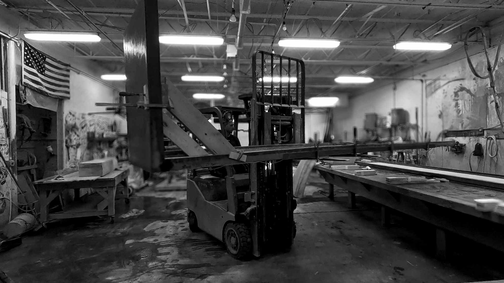 Black-and-white photograph of a forklift inside a concrete fabrication workshop, with long casting tables, overhead fluorescent lights, and raw industrial tools visible.