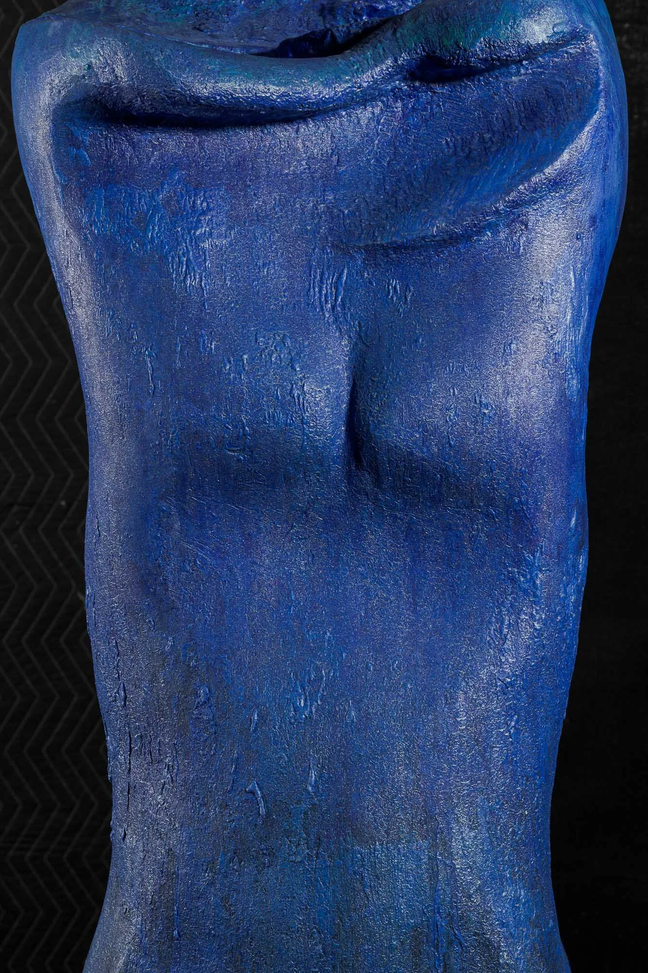 Close-up of a blue, textured sculpture of a human torso with defined abs and a black background.