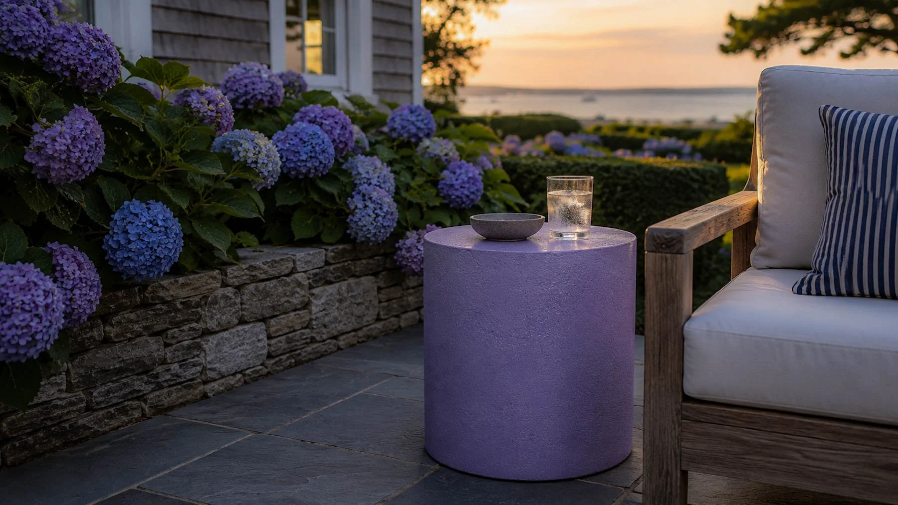 Concrete cylindrical side table positioned beside a lounge chair on a hydrangea-lined coastal terrace overlooking the water at sunset.