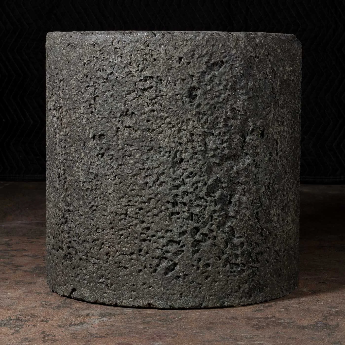 Dark charcoal concrete cylinder with porous volcanic surface texture and mineral pitting photographed in low light against a black backdrop.