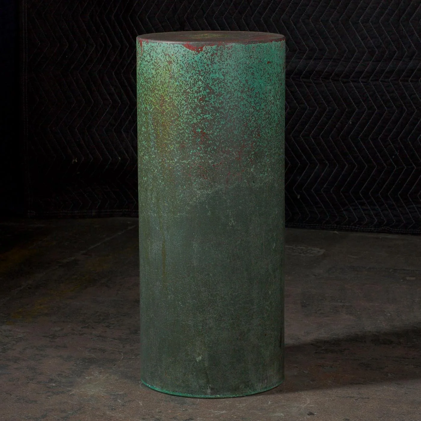 Tall cylindrical concrete form with oxidized copper-green surface variation and dark atmospheric finish photographed in studio