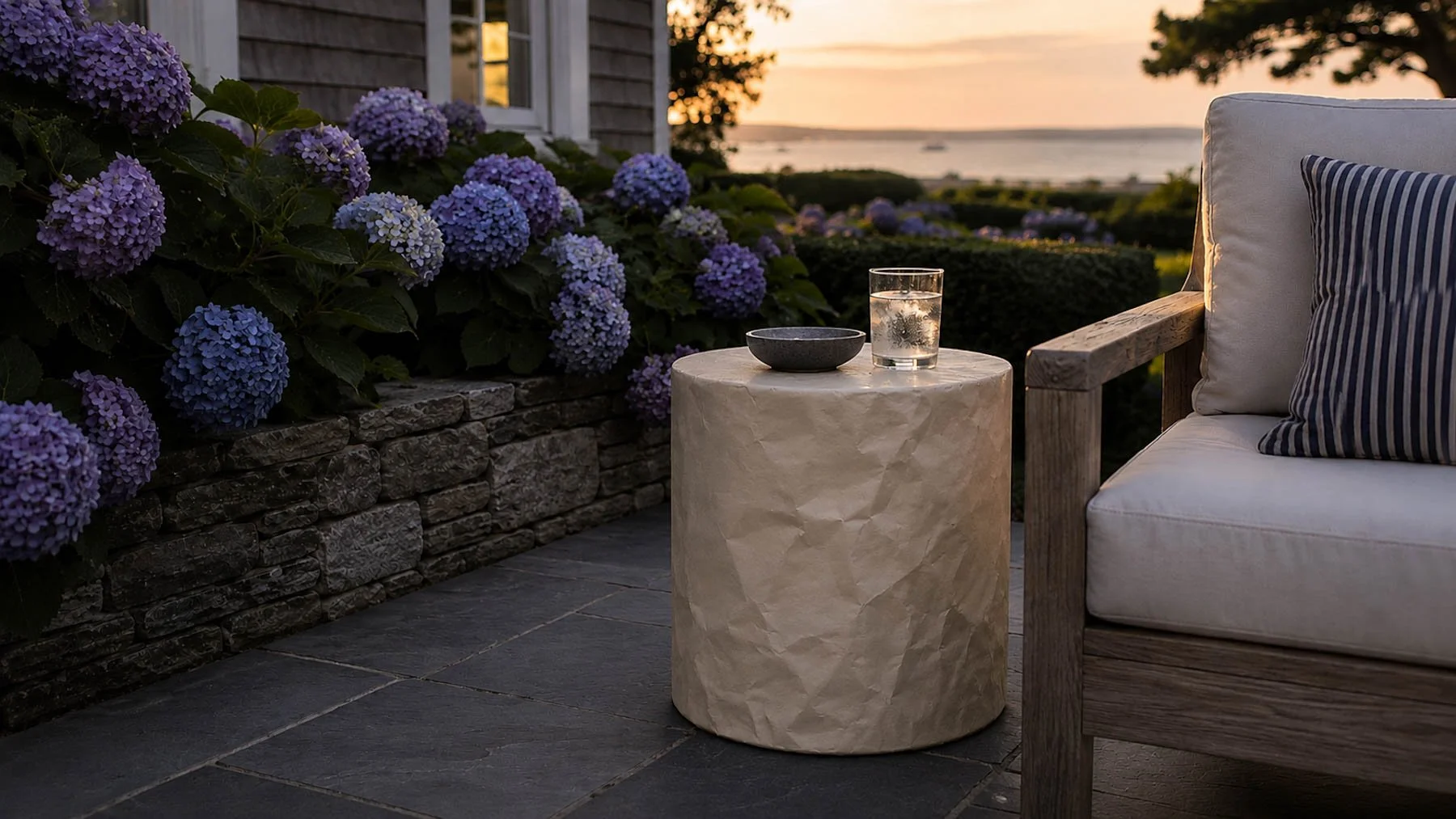 Paper-wrapped cylindrical form positioned beside a lounge chair on a hydrangea-lined coastal terrace overlooking the water at sunset. action:wrapped