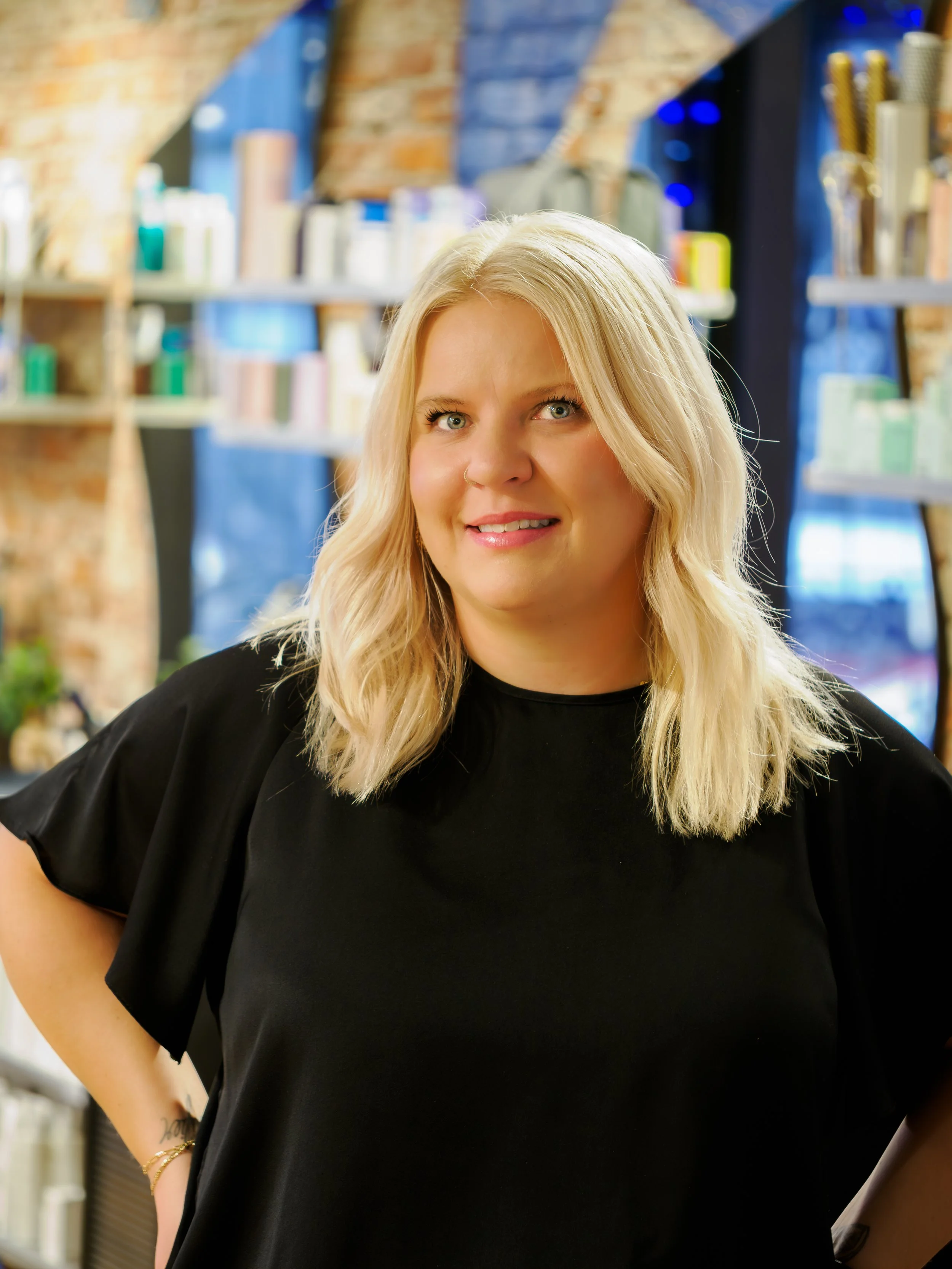 A woman with long, wavy auburn hair seen from behind, standing against a pink wall, with her hands resting on her head.