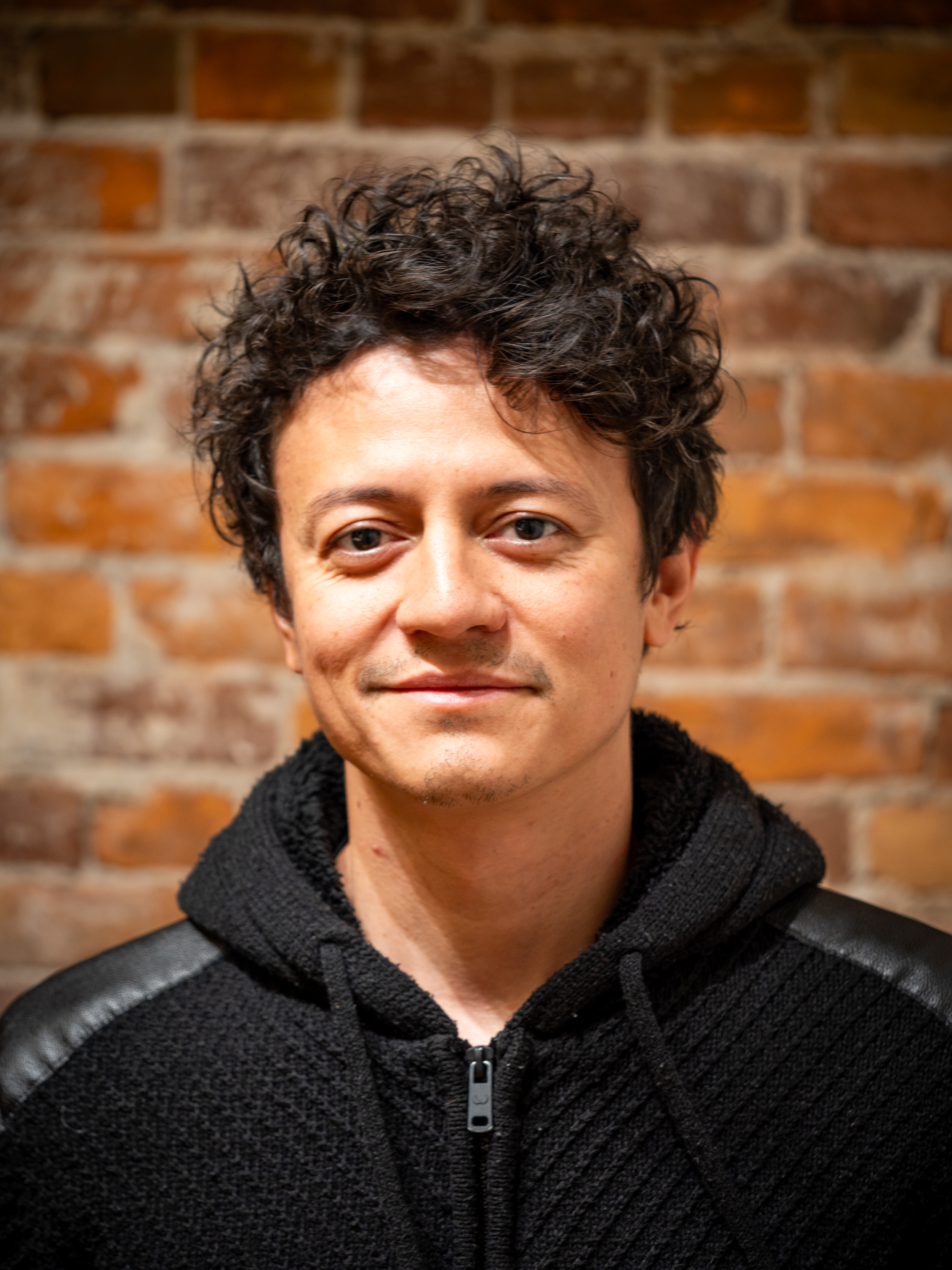 Headshot of a man with curly dark hair, wearing a black textured jacket with leather details on the shoulders, standing against a brick wall background.