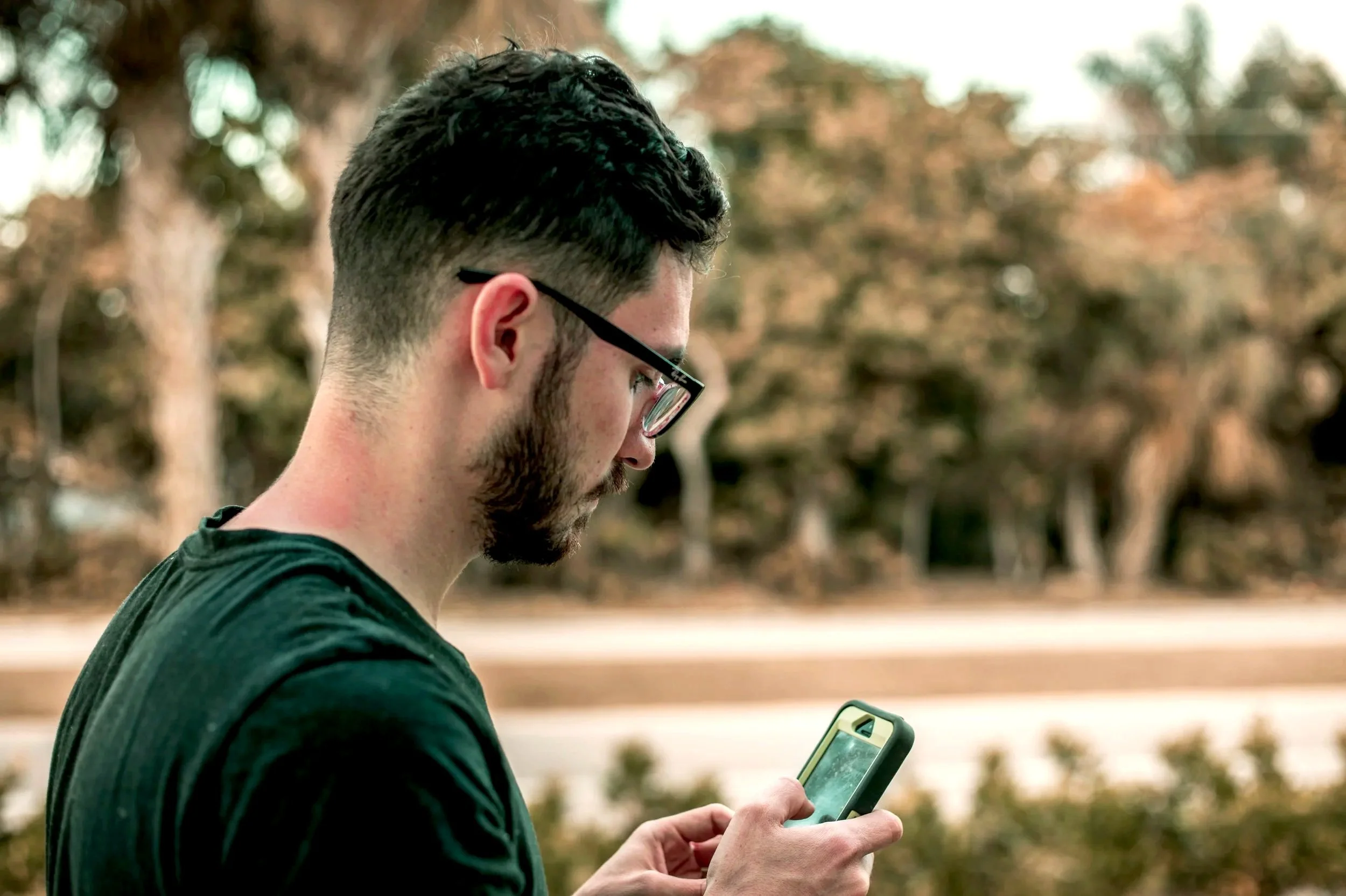 A man with dark hair, glasses, and a beard looks at his phone outdoors with a fall-colored forest in the background. He's logging his meals for the day to see his macronutrients and micronutrients.