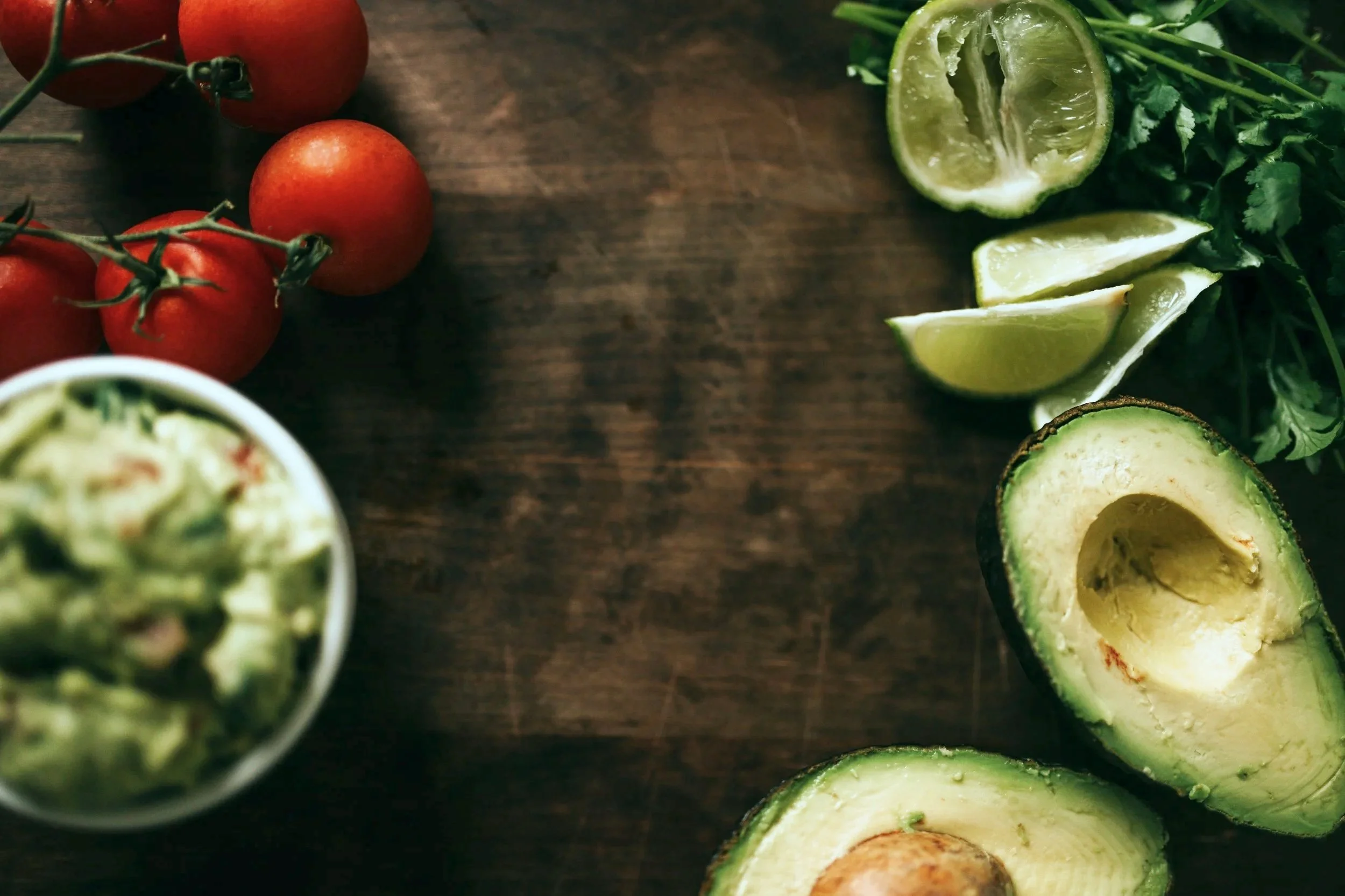 Tomatoes on a vine, lime wedges, avocado, fresh cilantro, and a bowl of guacamole on a wooden surface. These are the ingredients that make up the healthy meal you will track in the Nutrition Ambition app.