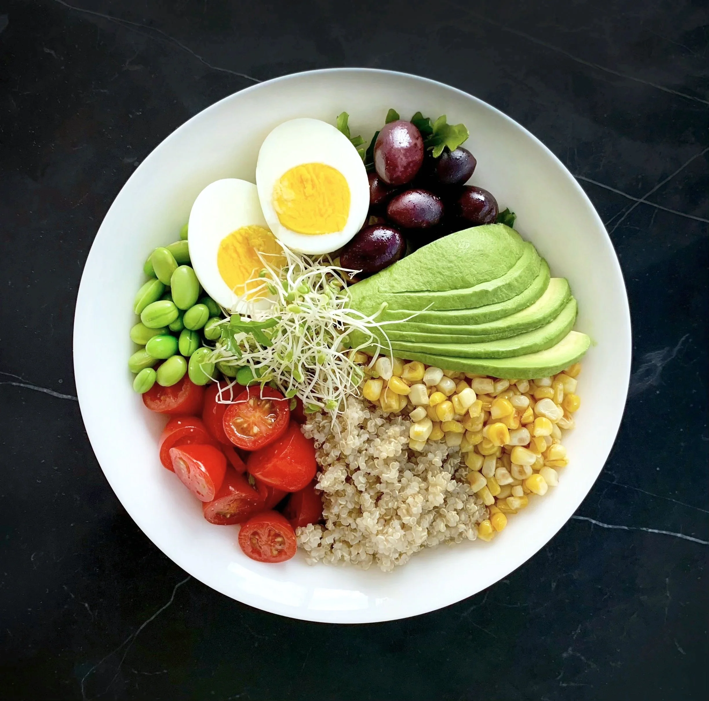 A white bowl filled with sliced avocado, cherry tomatoes, cooked quinoa, corn, alfalfa sprouts, olives, halved boiled eggs, edamame beans, and halved cherry tomatoes on a dark surface. Log this meal with Nutrition Ambition to see its nutrients.
