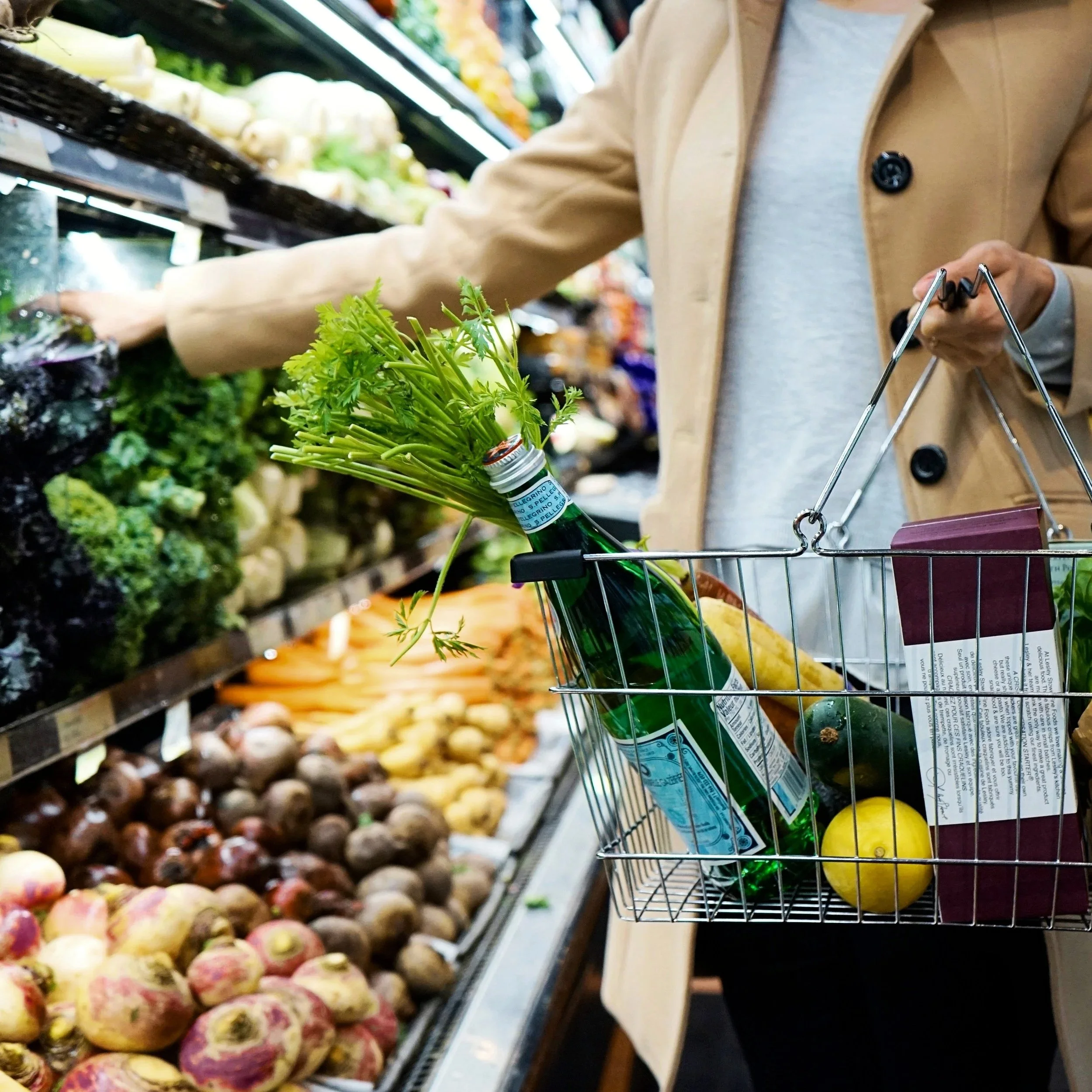 A shopper in a supermarket holds a small shopping basket containing a bottle with leafy greens, a yellow lemon, and a purple packaged item, near a produce display with various vegetables. Use Nutrition Ambition to help you shop for healthy choices.