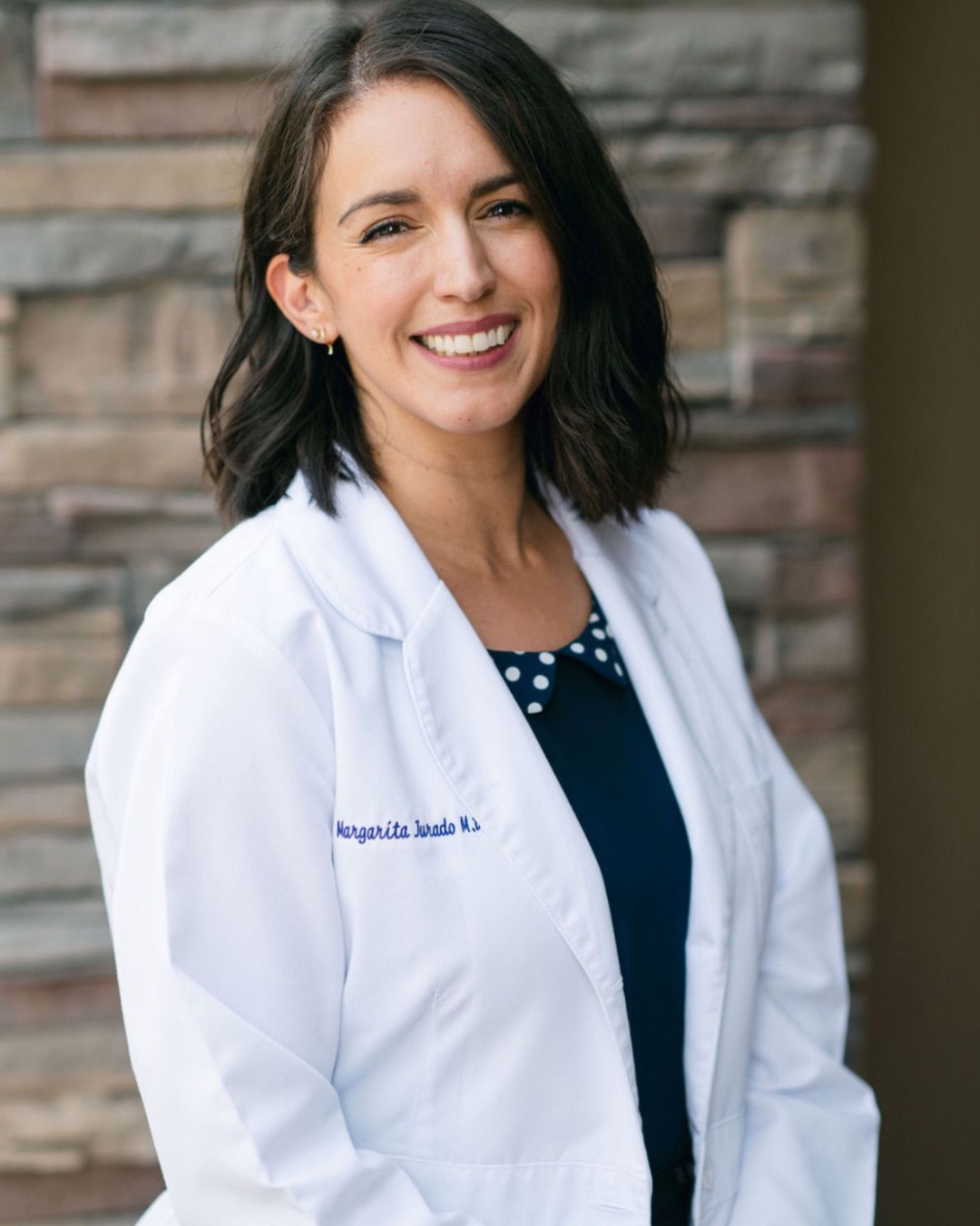 Smiling female doctor with a short haircut, wearing a white lab coat over a navy blouse with a polka-dot collar, accessorized with small gold earrings, standing in front of a stone wall background. Kern Place Pediatrics.