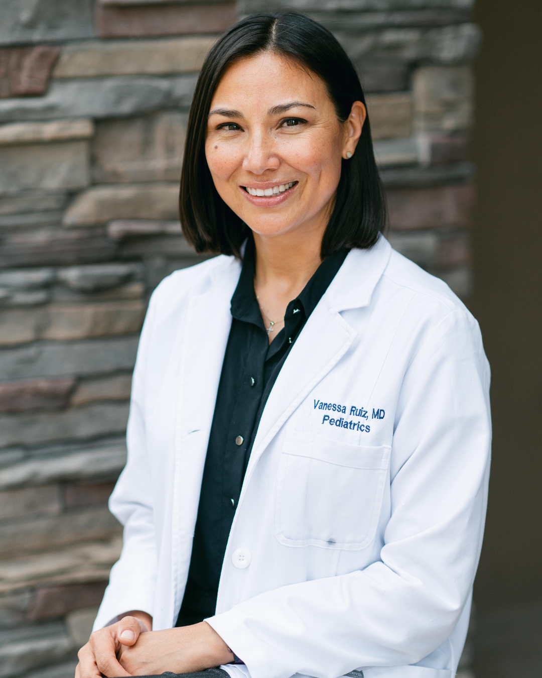Smiling female doctor wearing a white lab coat over a black shirt, standing in front of a stone wall background. Kern Place Pediatrcis.