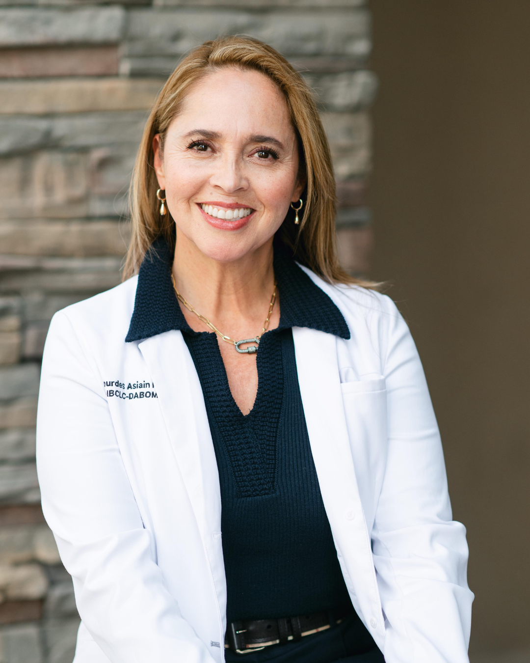 miling female doctor wearing a white lab coat over a dark navy top, accessorized with small hoop earrings and layered gold necklaces, sitting in front of a stone wall background. Kern Place Pediatrics.