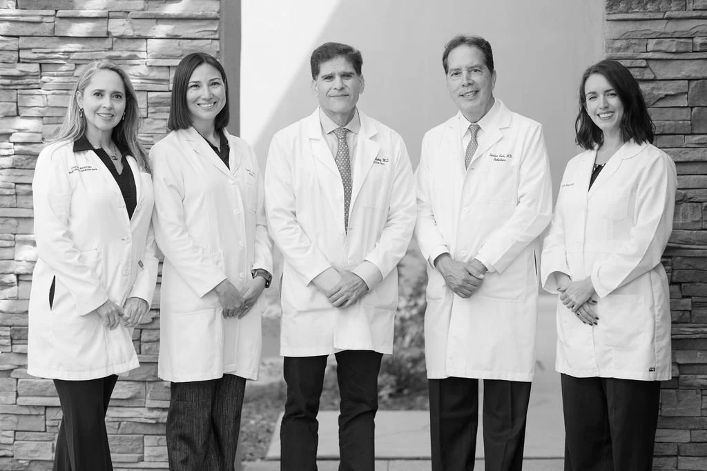 Black and white group photo of five doctors wearing white lab coats, standing outdoors in front of a stone wall and smiling at the camera. Two men and three women are posed together in a professional setting.