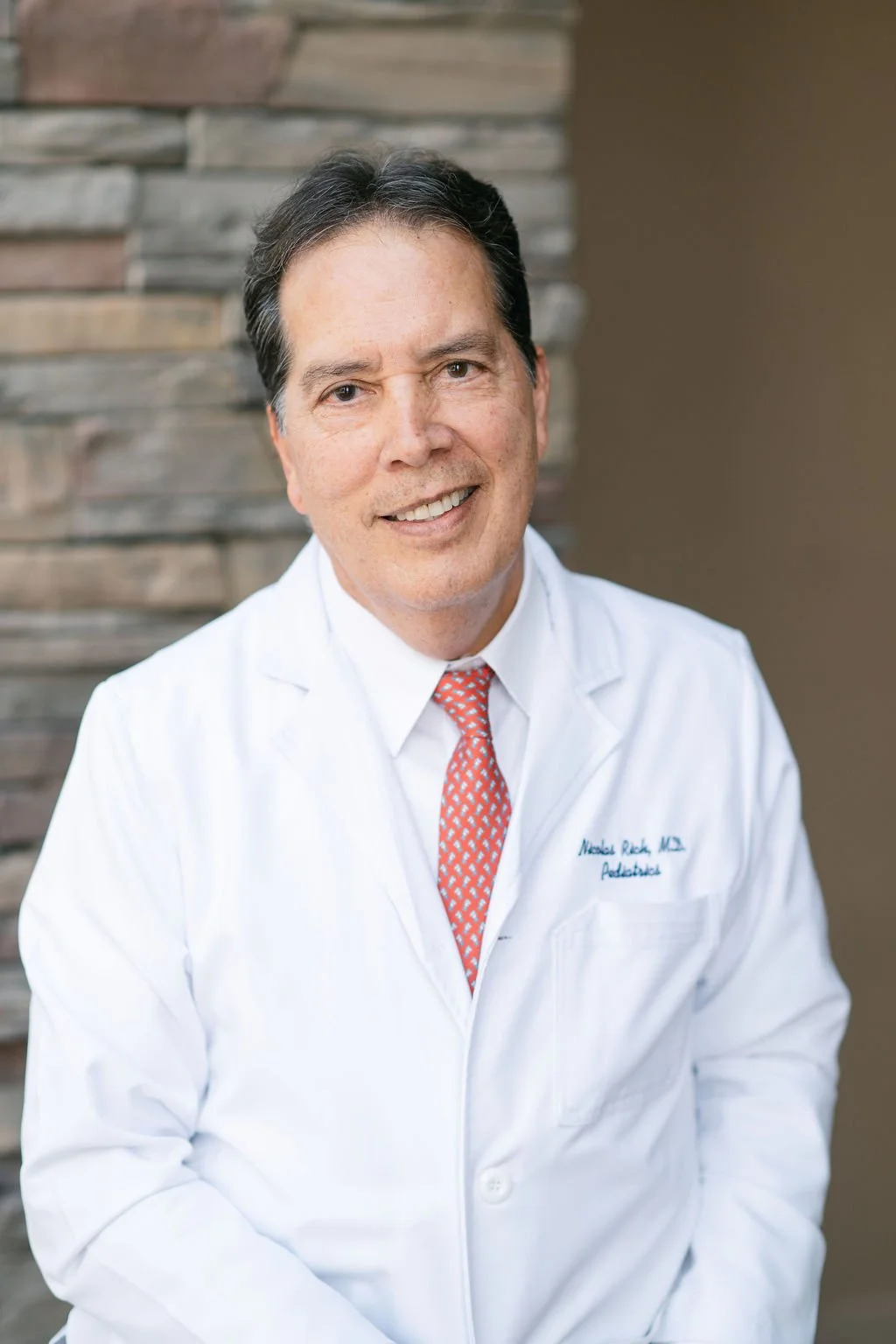 Smiling male doctor wearing a white lab coat and red patterned tie, standing in front of a stone wall background. Kern Place Pediatrcis.