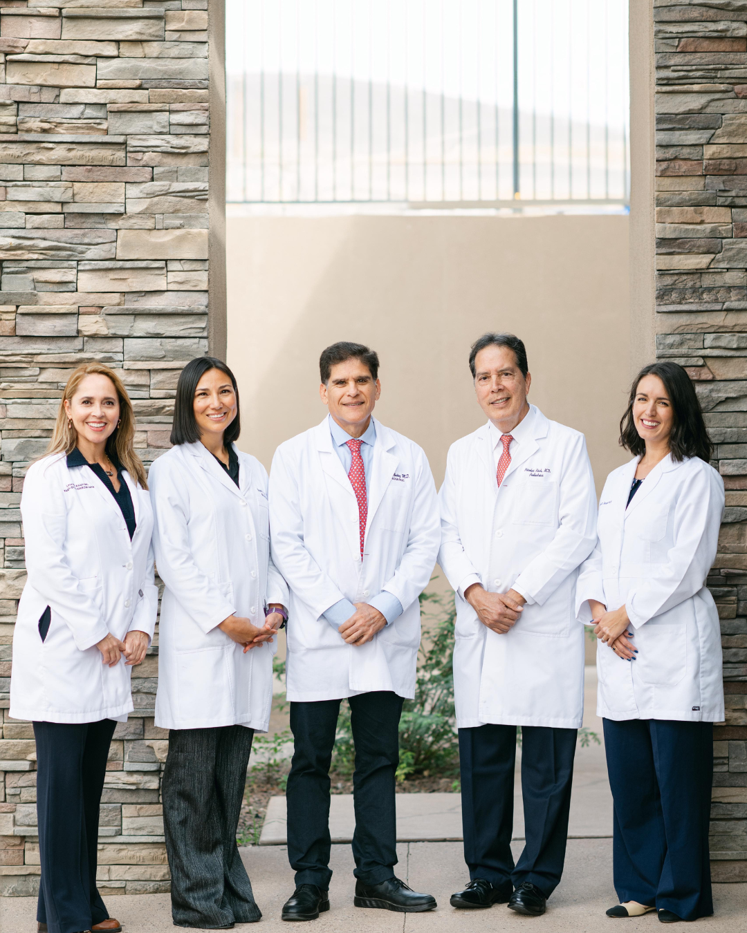 Group of five doctors wearing white lab coats, standing outdoors in front of a stone wall and smiling at the camera. Two men and three women are posed together, appearing professional and friendly.