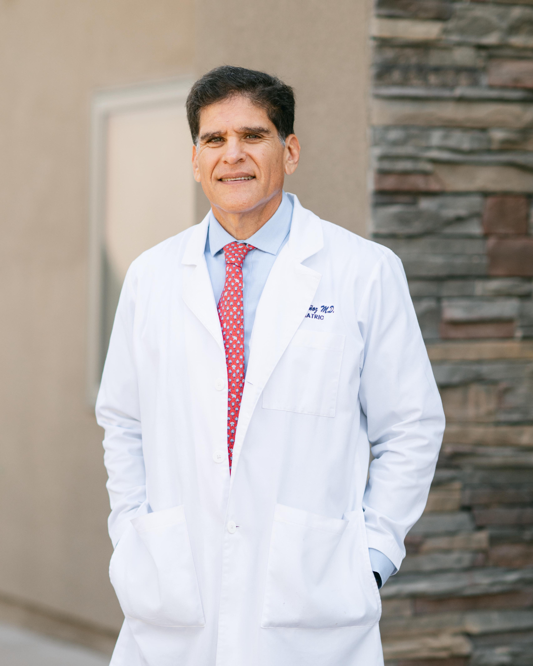 Male doctor wearing a white lab coat, blue shirt, and red patterned tie, standing outdoors in front of a stone and beige wall, smiling slightly.