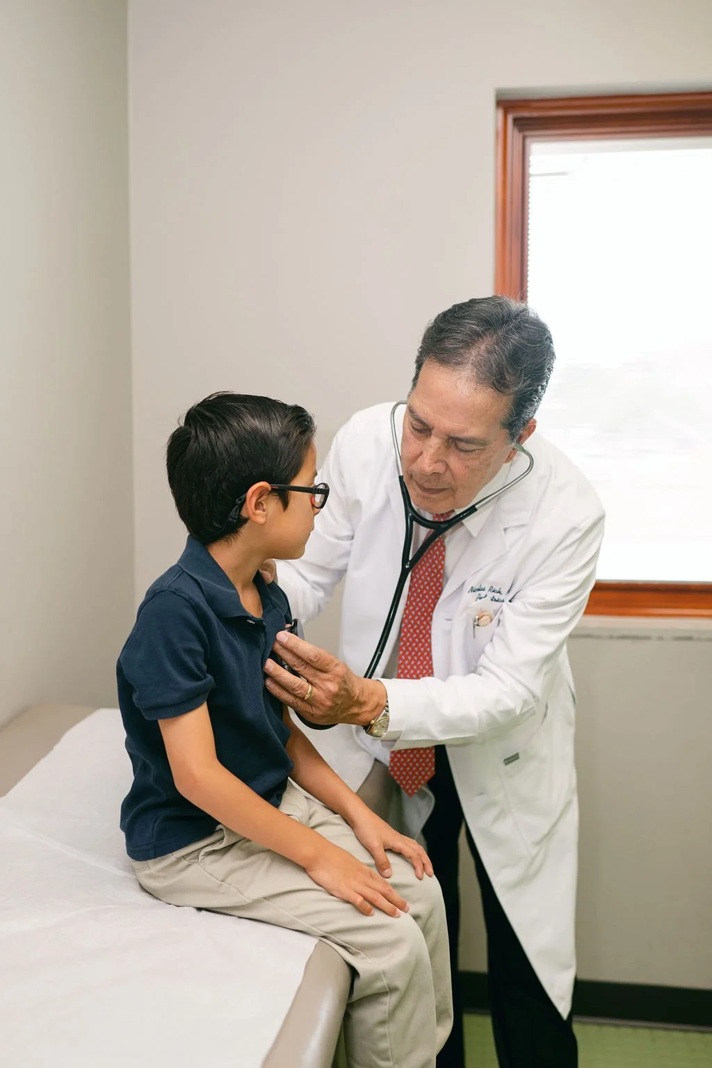 Male doctor wearing a white lab coat using a stethoscope to examine a young boy sitting on an exam table in a medical office. Kern Place Pediatrics.
