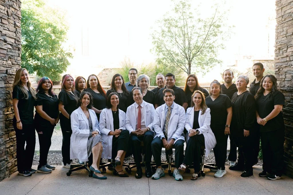 A group photo of a medical team standing and sittin outdoors between two stone walls. Three doctors in white coats sit in front, surrounded by staff members dressed in black scrubs. The group is smillin and posing together in natural light with trees