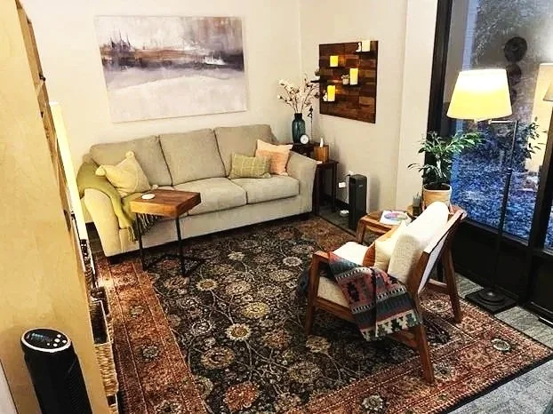 Living room with beige sofa, two armchairs, a patterned rug, a large window, a floor lamp, houseplants, wall art, and side tables.