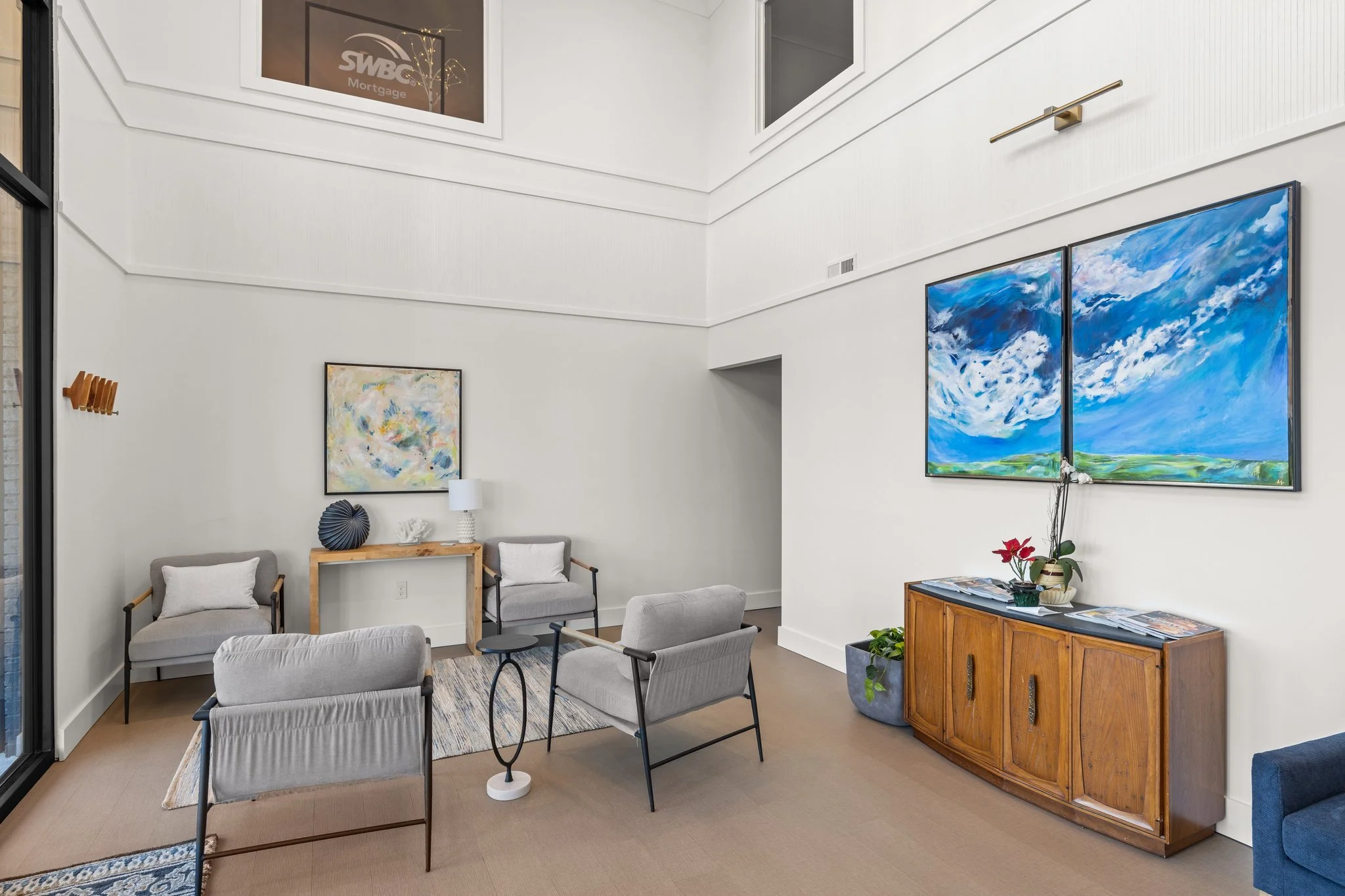 Modern lobby or waiting area with gray chairs, a wooden cabinet, colorful paintings, and a glass door.
