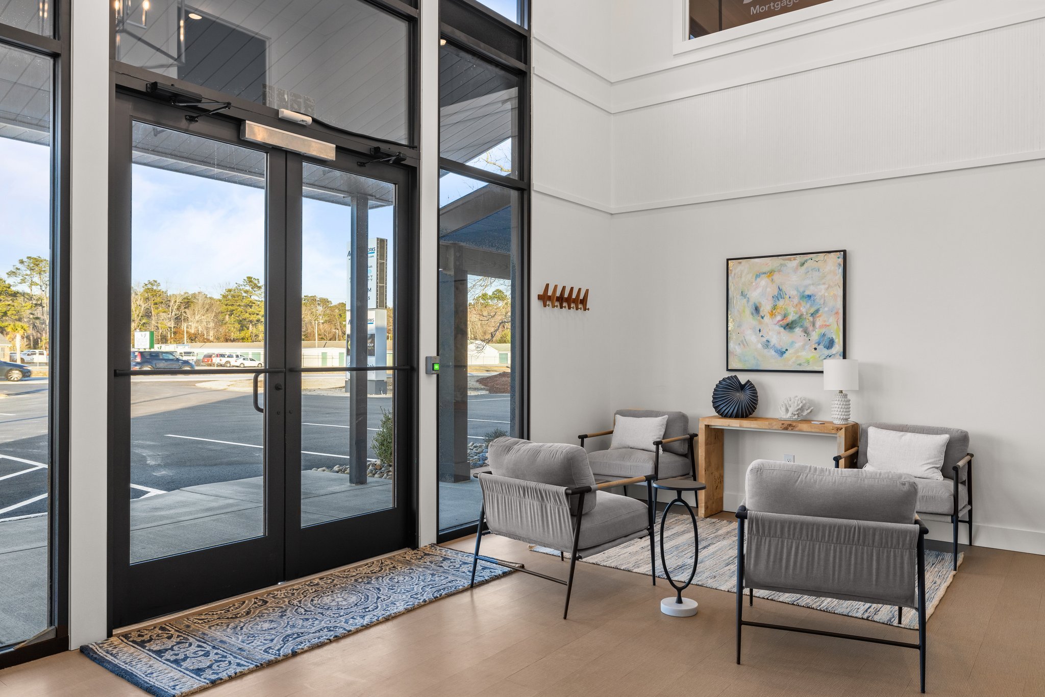 Cozy lobby with gray armchairs, a wooden side table, a white table lamp, a colorful abstract art piece, and a decorative black vase. Large windows and glass doors with a view of Morehead City's Highway 70