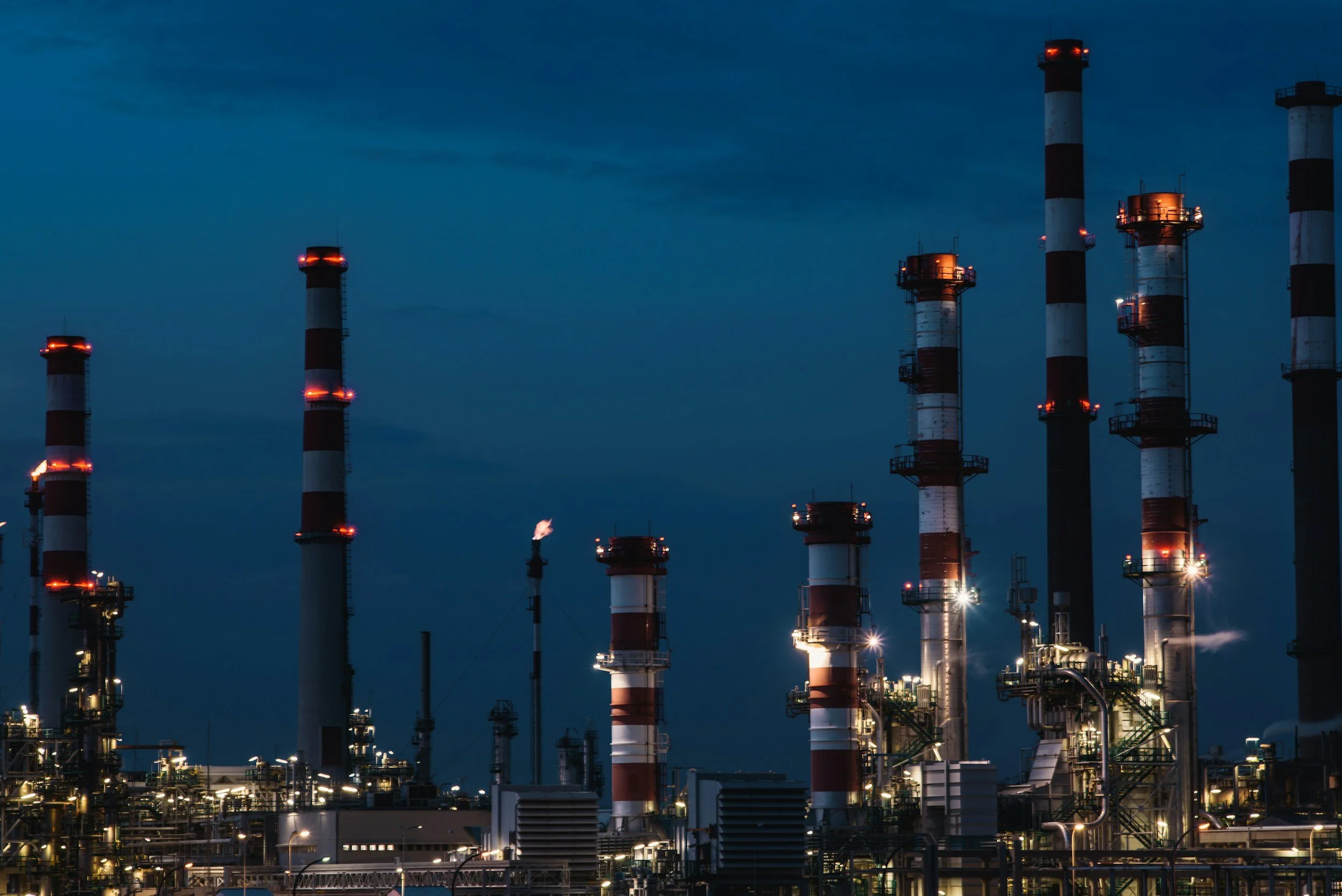 Night view of an industrial oil refinery with several tall smokestacks emitting no visible smoke, illuminated by bright industrial lights.