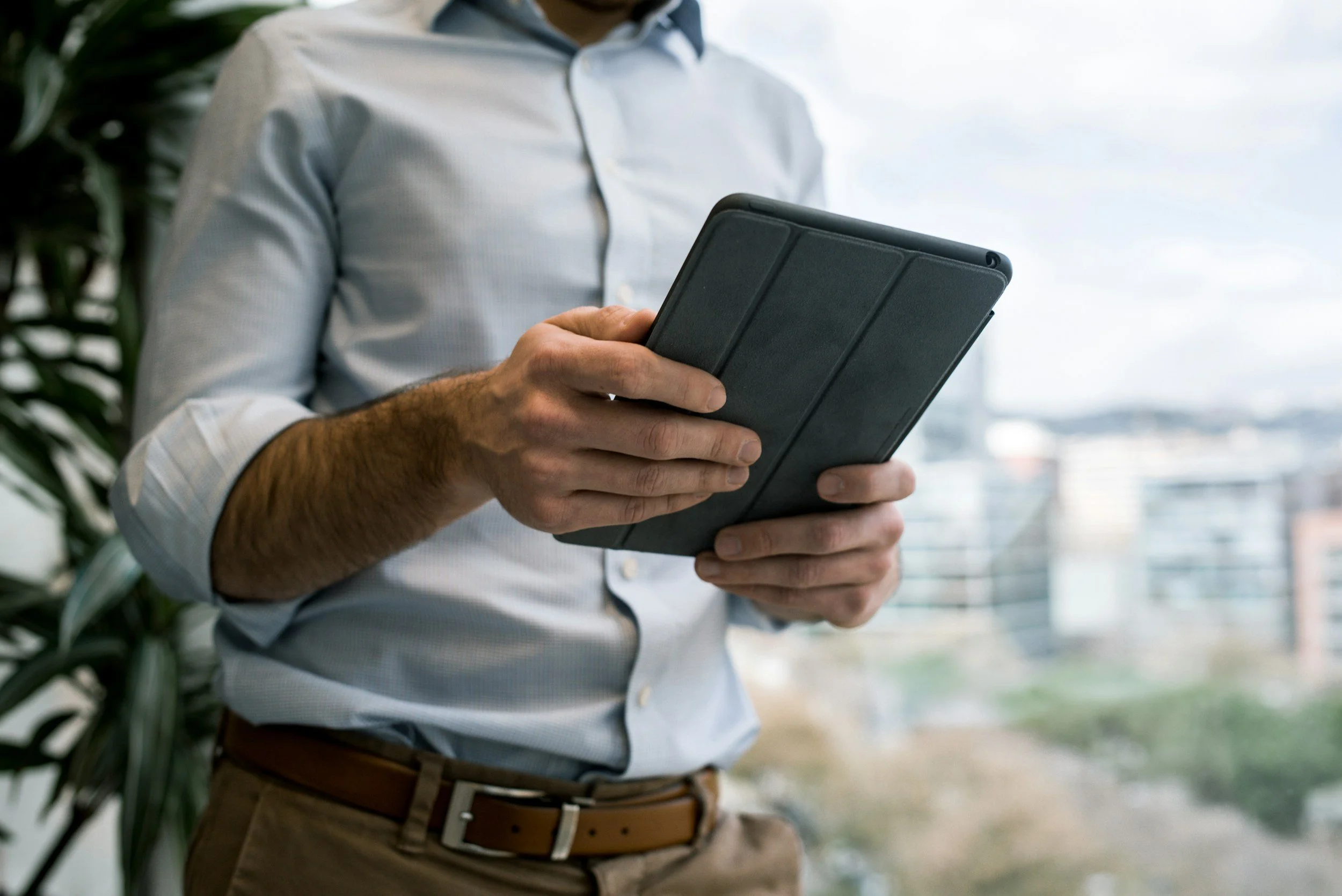 A man in a light blue shirt holding and looking at a black tablet in an office or urban setting.