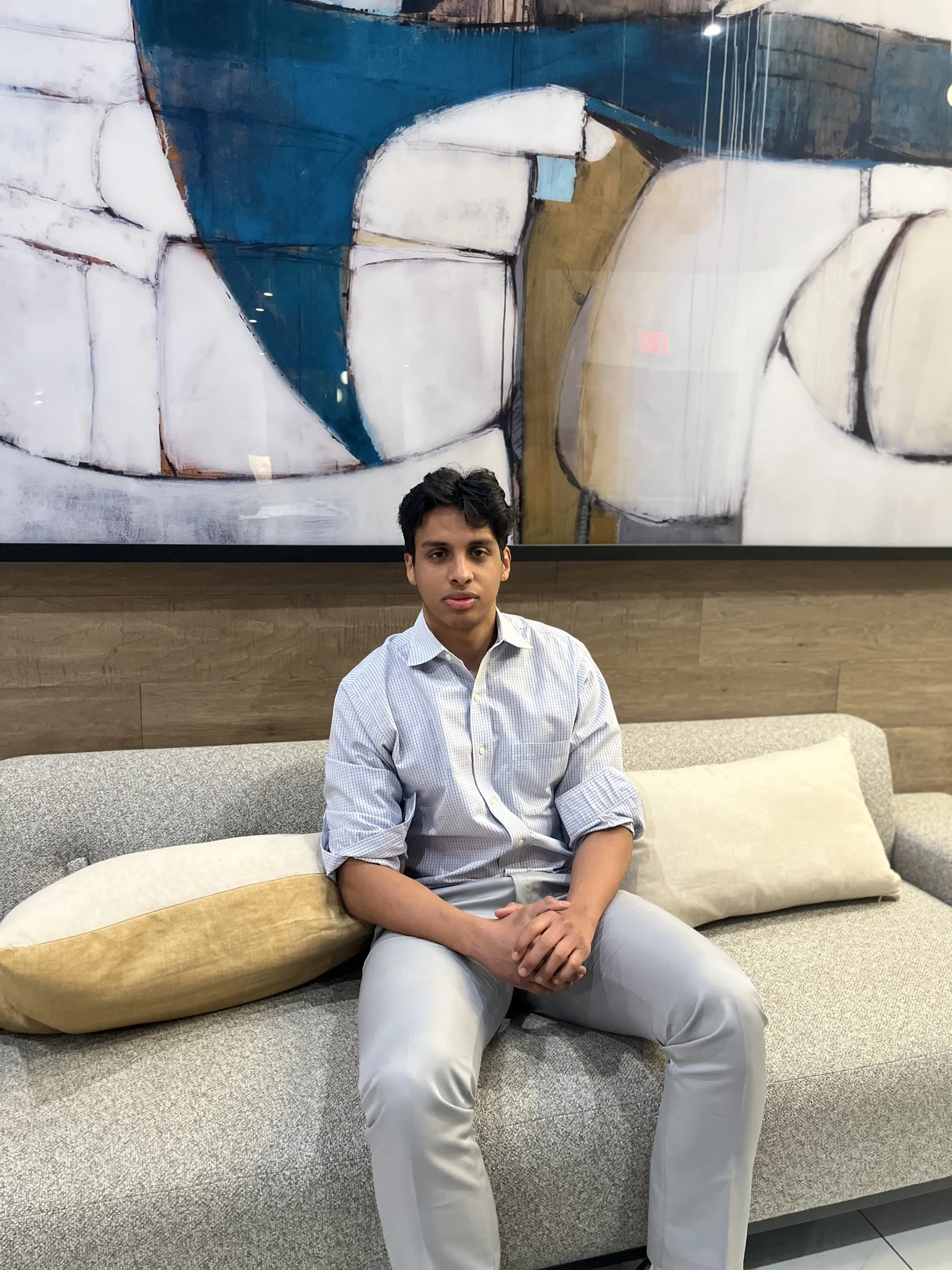 A young man with glasses sitting on a teal couch, wearing a dark gray zip-up sweater and light blue pants, with a beige and white textured pillow beside him.