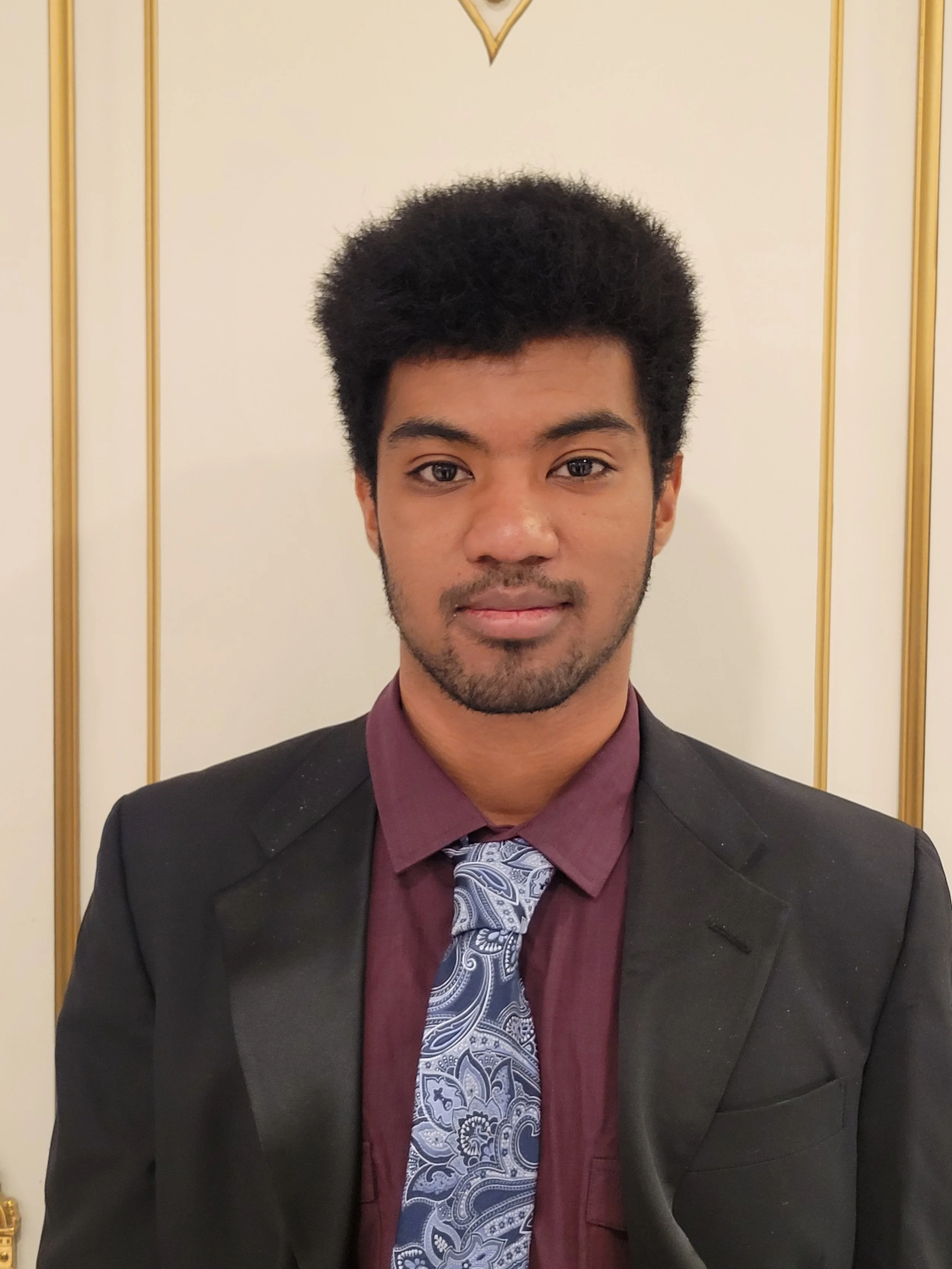 A young man with dark curly hair, wearing a dark suit jacket, maroon shirt, and a patterned tie, standing in front of a light-colored wall with gold accents.