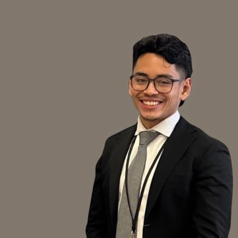 A young man in a black suit, white shirt, and gray tie smiling at the camera, wearing glasses, against a plain background.