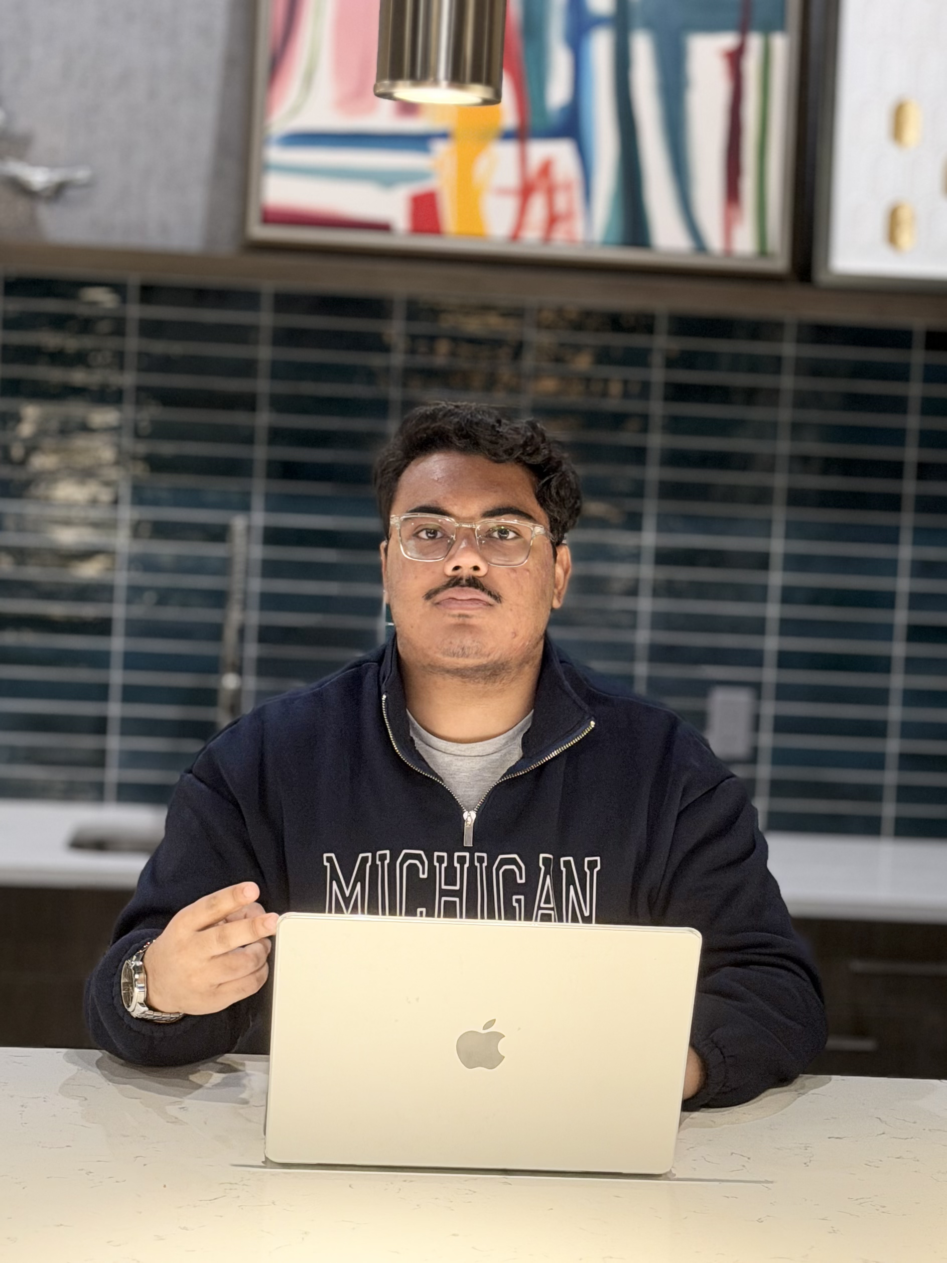 A man with glasses and a mustache sitting at a table with an open MacBook, pointing at the laptop in a modern indoor space.