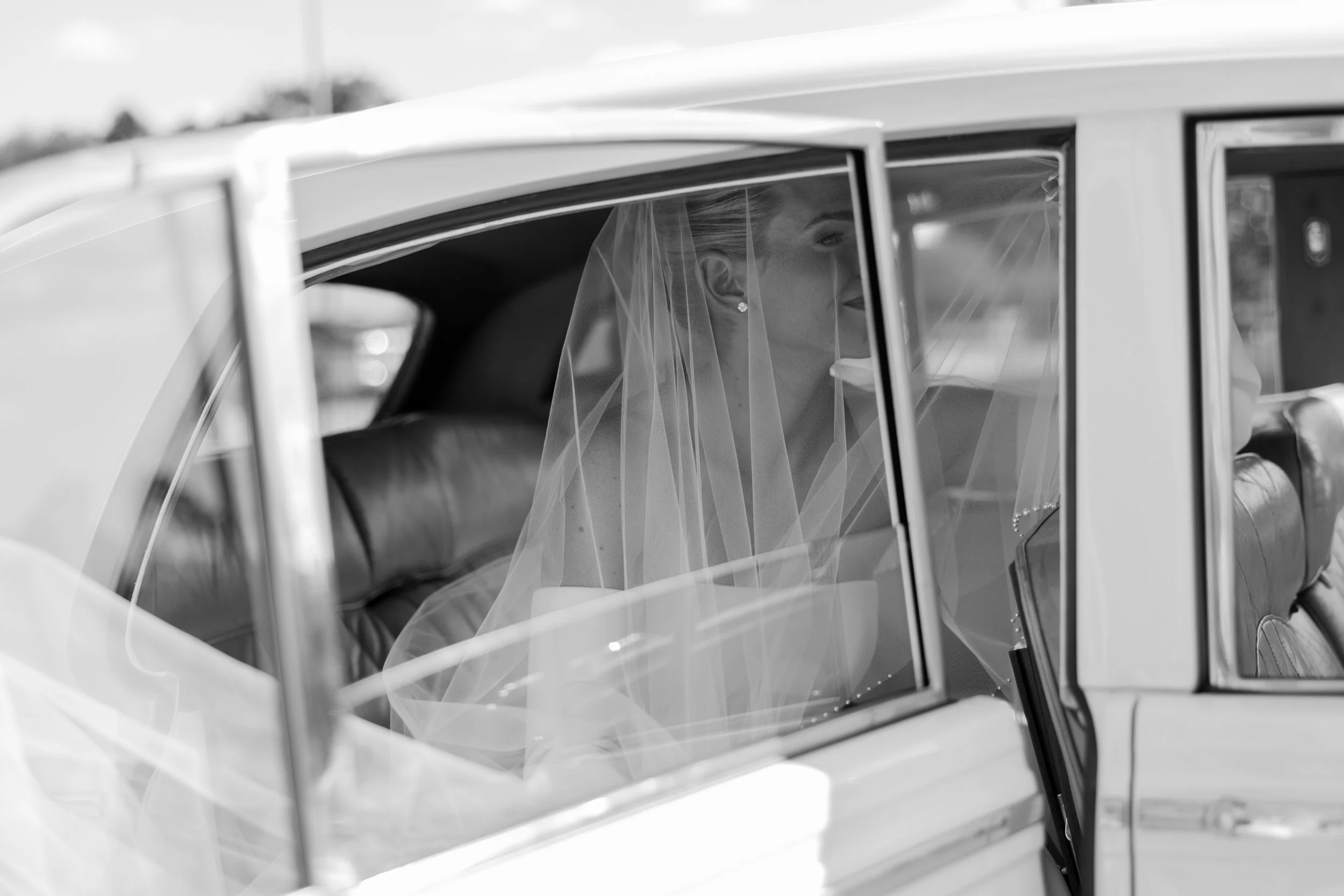 A bride sitting inside a vintage car, wearing a veil and elegant earrings, with her face turned to the side and smiling.