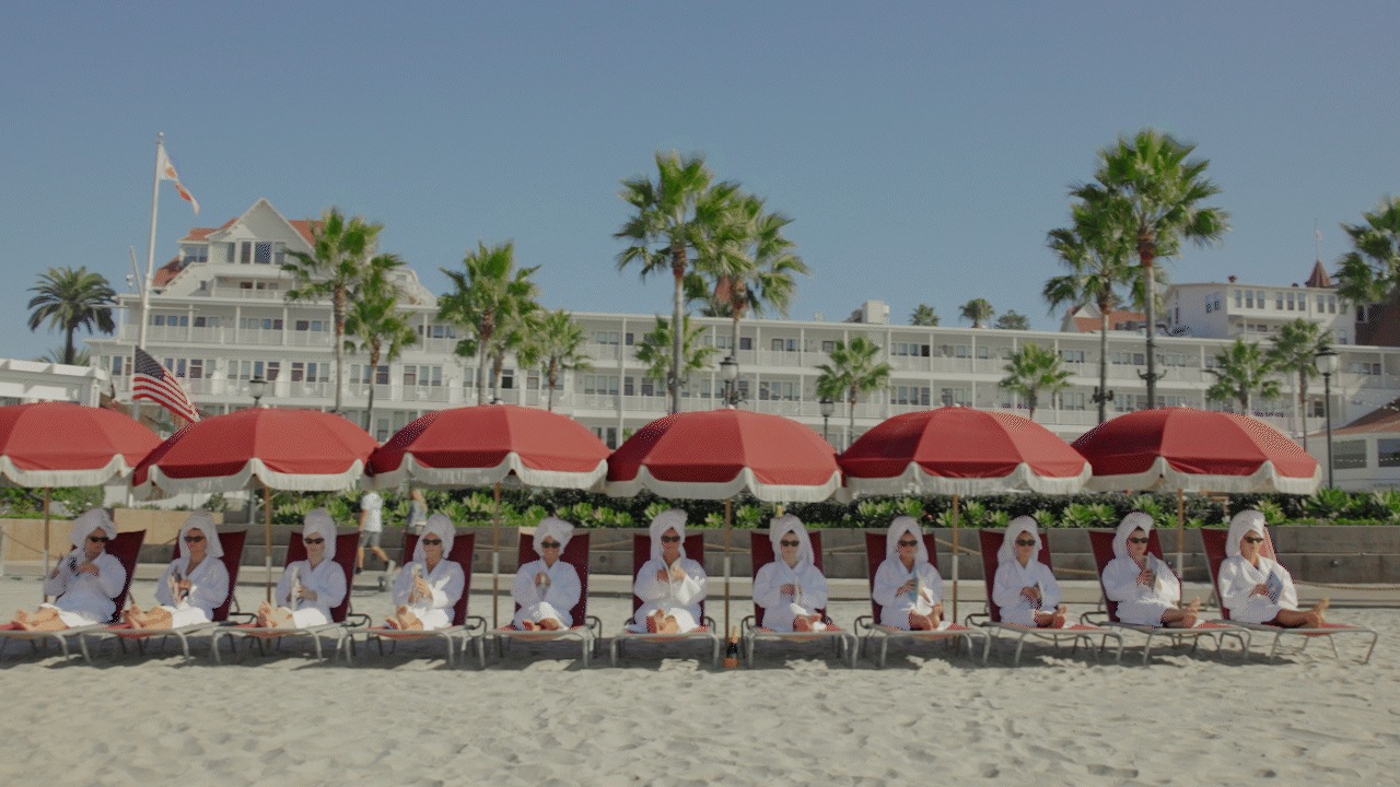 Multiple women wearing white robes and sunglasses are sitting on lounge chairs under red umbrellas on a beach, with a building and palm trees in the background.