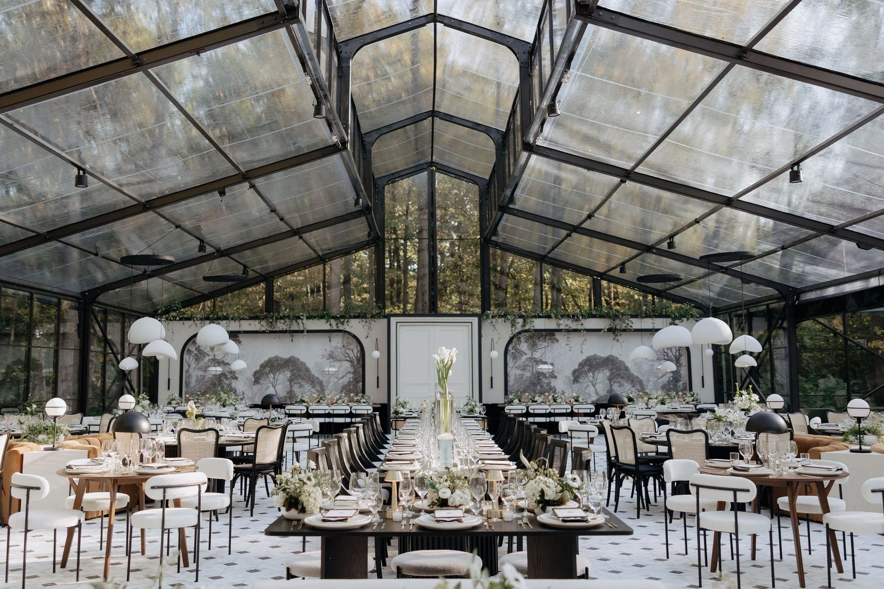 Elegant indoor event space with a high glass ceiling, decorated with white and black accents, floral arrangements, and modern lighting fixtures.