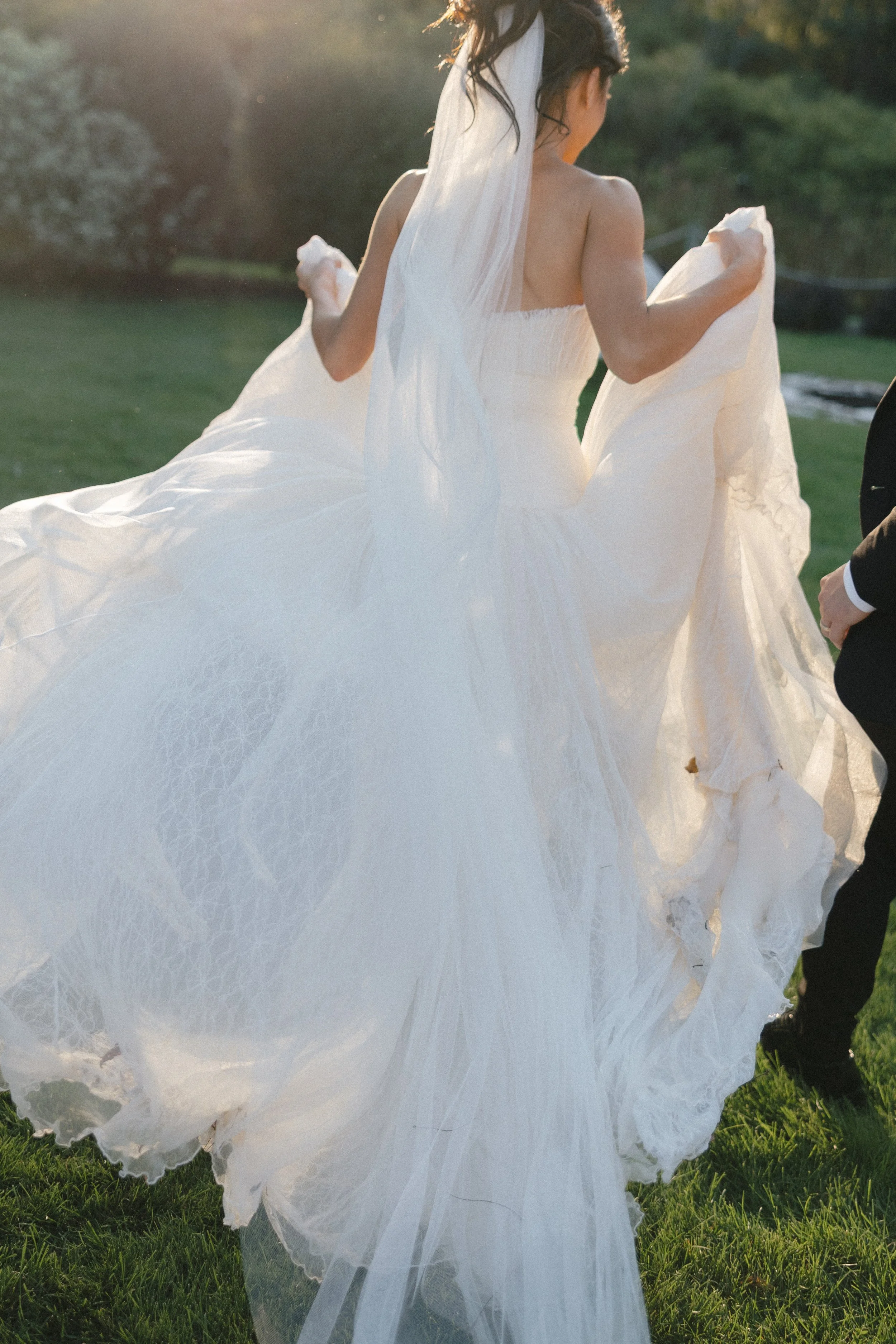 A bride in a flowing white wedding dress outdoors, holding up the skirt as she walks on grass, with a groom partially visible on the right.