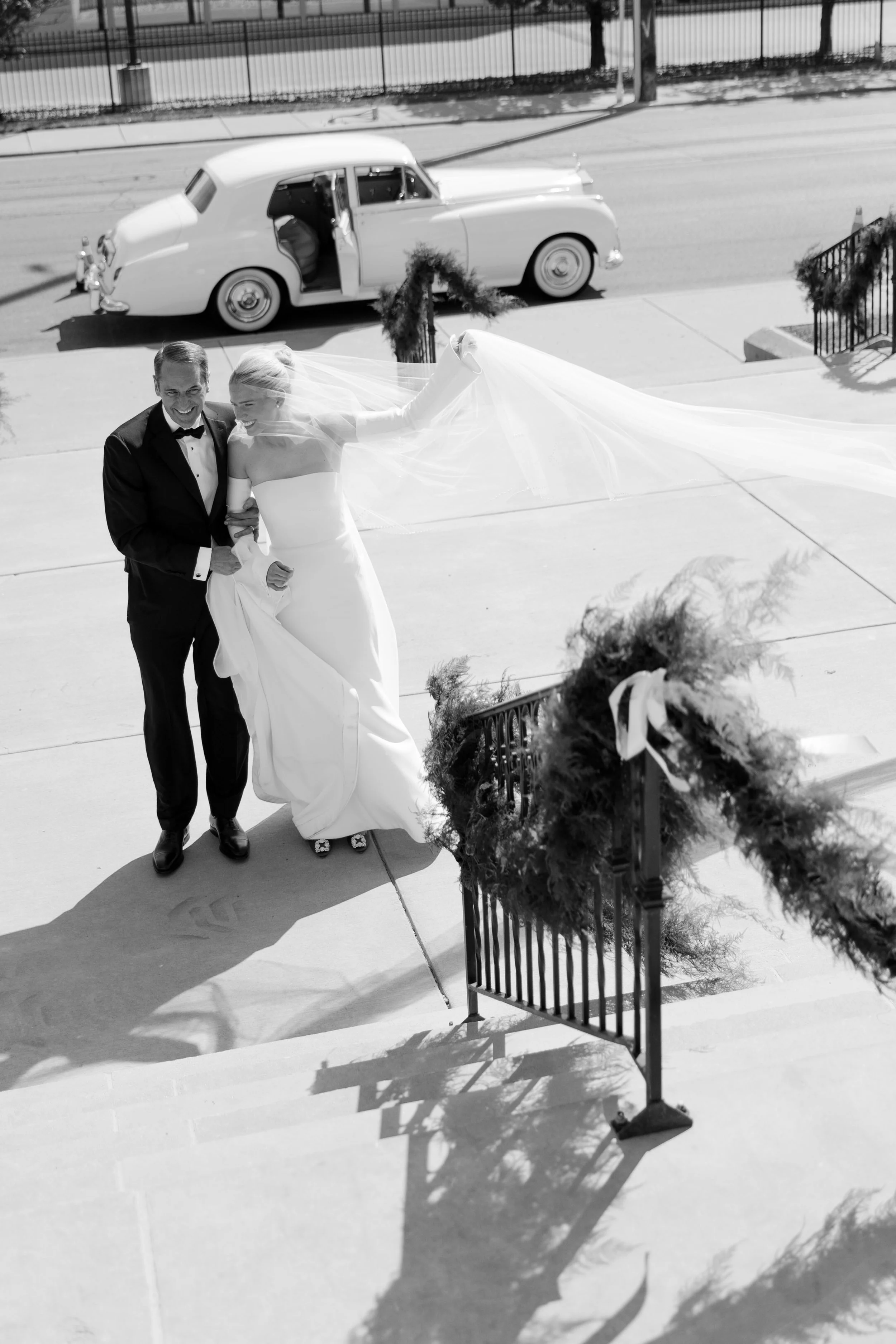 A bride in a white wedding gown and a groom in a tuxedo walking up steps outside a building, with a vintage car parked on the street behind them.