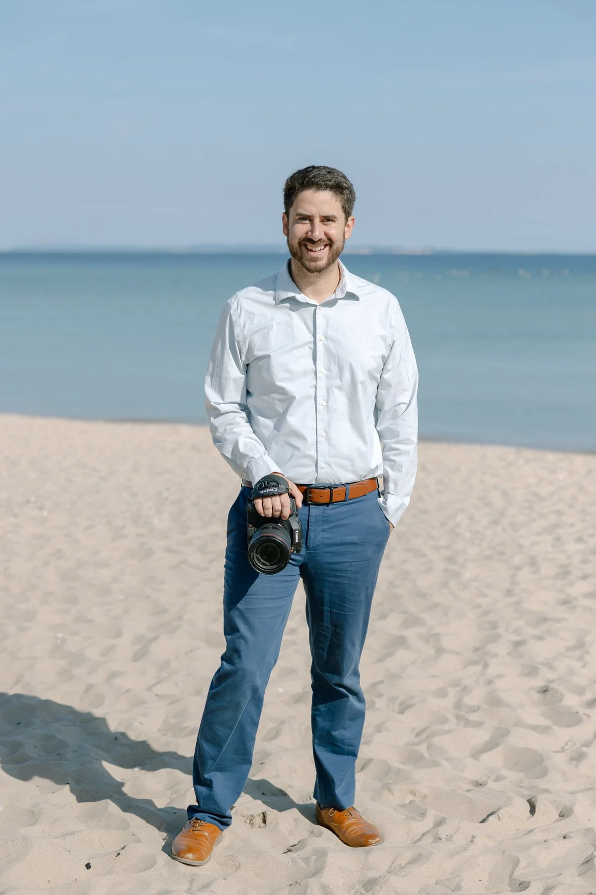 A man in a white shirt and blue pants standing on a sandy beach, holding a professional camera, with the ocean and a blue sky in the background.