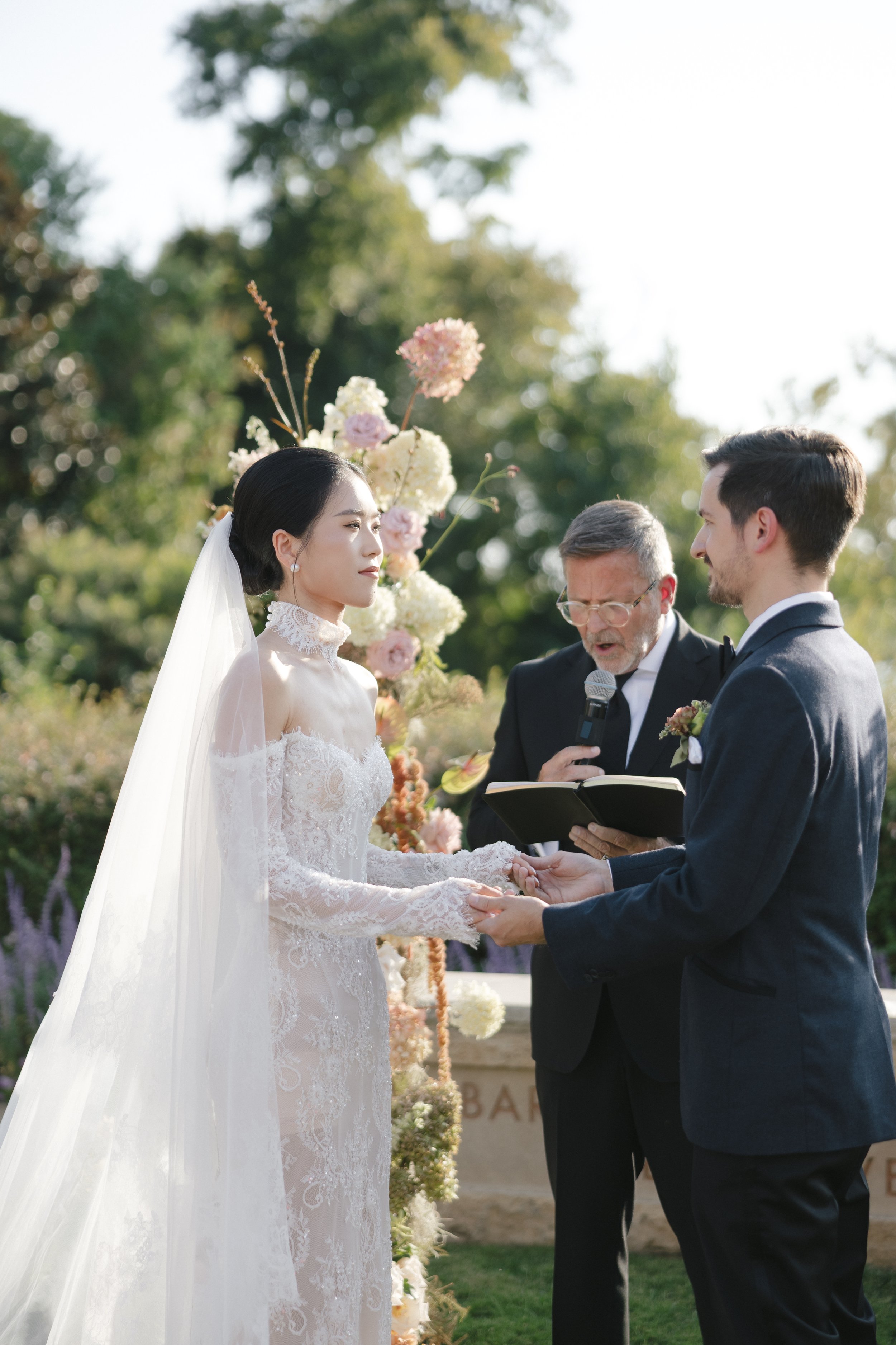 A couple getting married outdoors, holding hands and facing each other, with an officiant reading from a book. The bride is in a lace wedding dress and veil, and the groom is in a navy suit. There are flowers and greenery in the background.