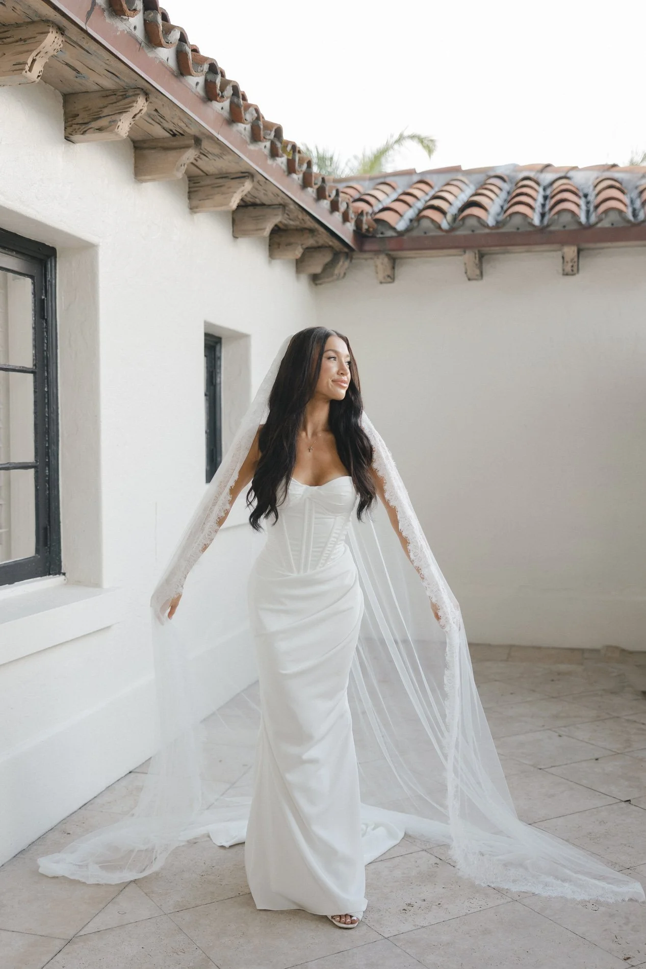 A bride in a white wedding gown with a long veil stands outdoors on a tiled patio in front of a white stucco wall with windows and a red-tiled roof. The bride has long dark hair and is looking to her right with a serene expression.