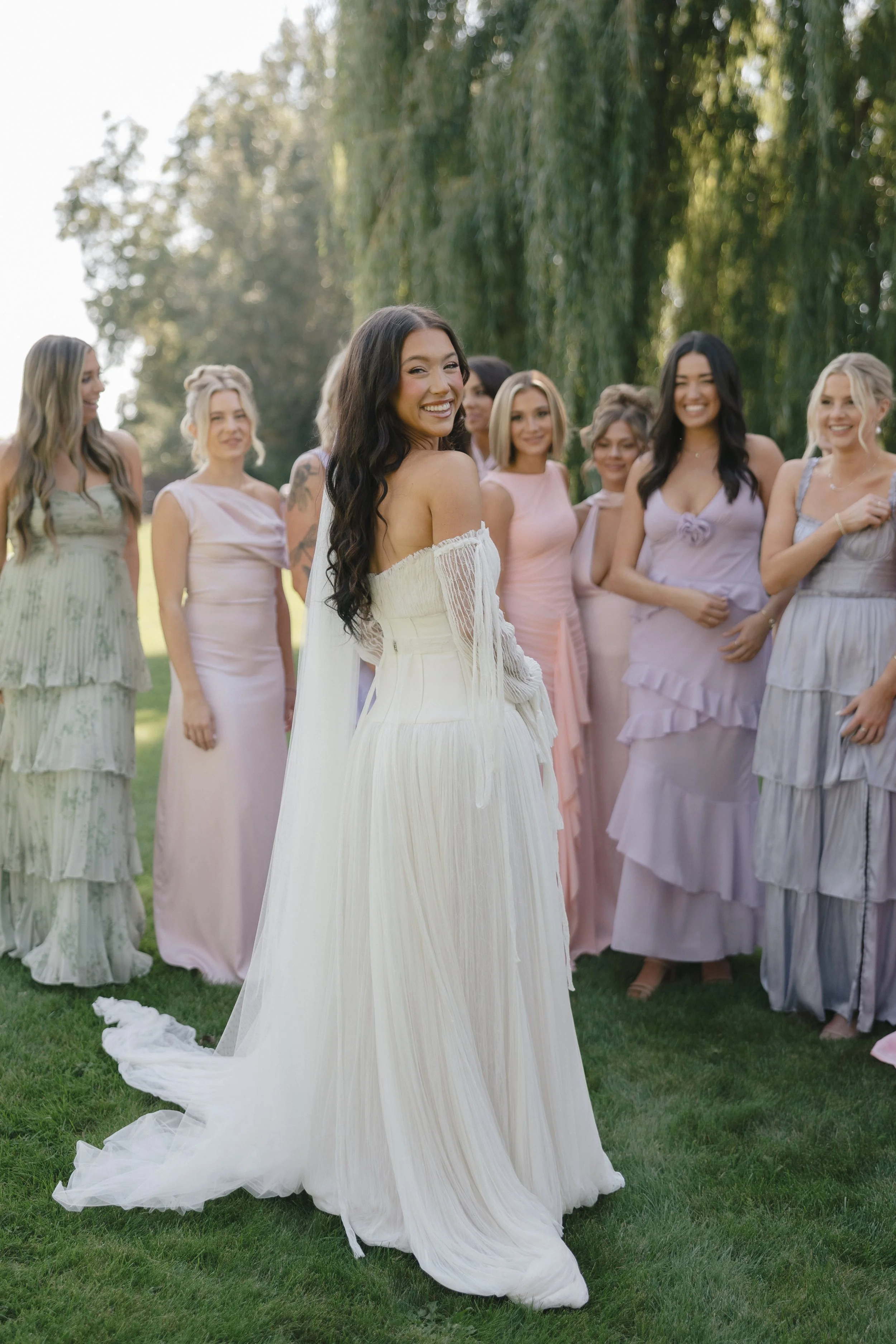 A bride in a white gown turns and smiles, surrounded by bridesmaids in pastel dresses, outdoors on a green lawn with trees in the background.