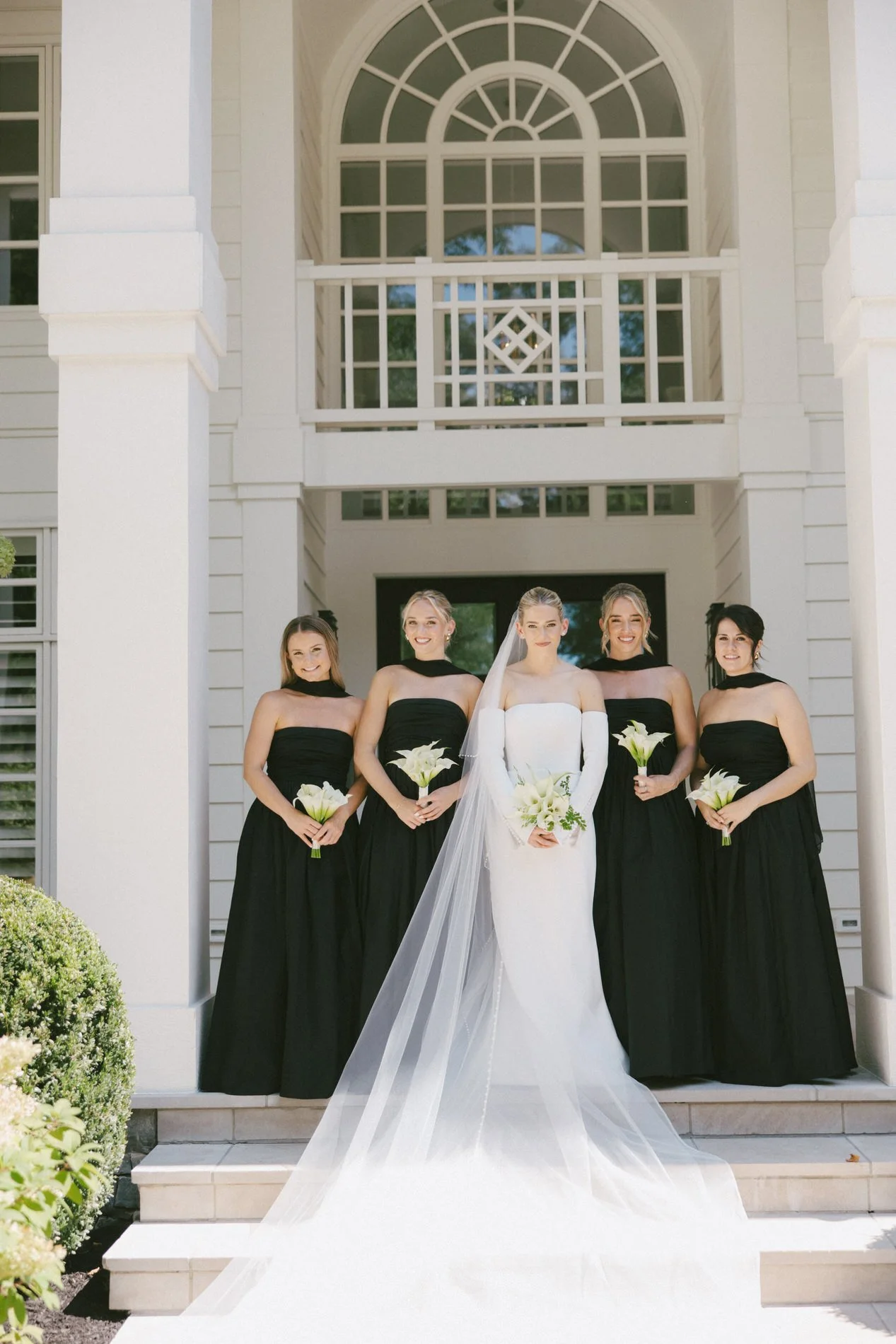 A bride in a white wedding gown and a veil, standing on steps in front of a building with large windows, surrounded by four bridesmaids dressed in black gowns holding white bouquets.