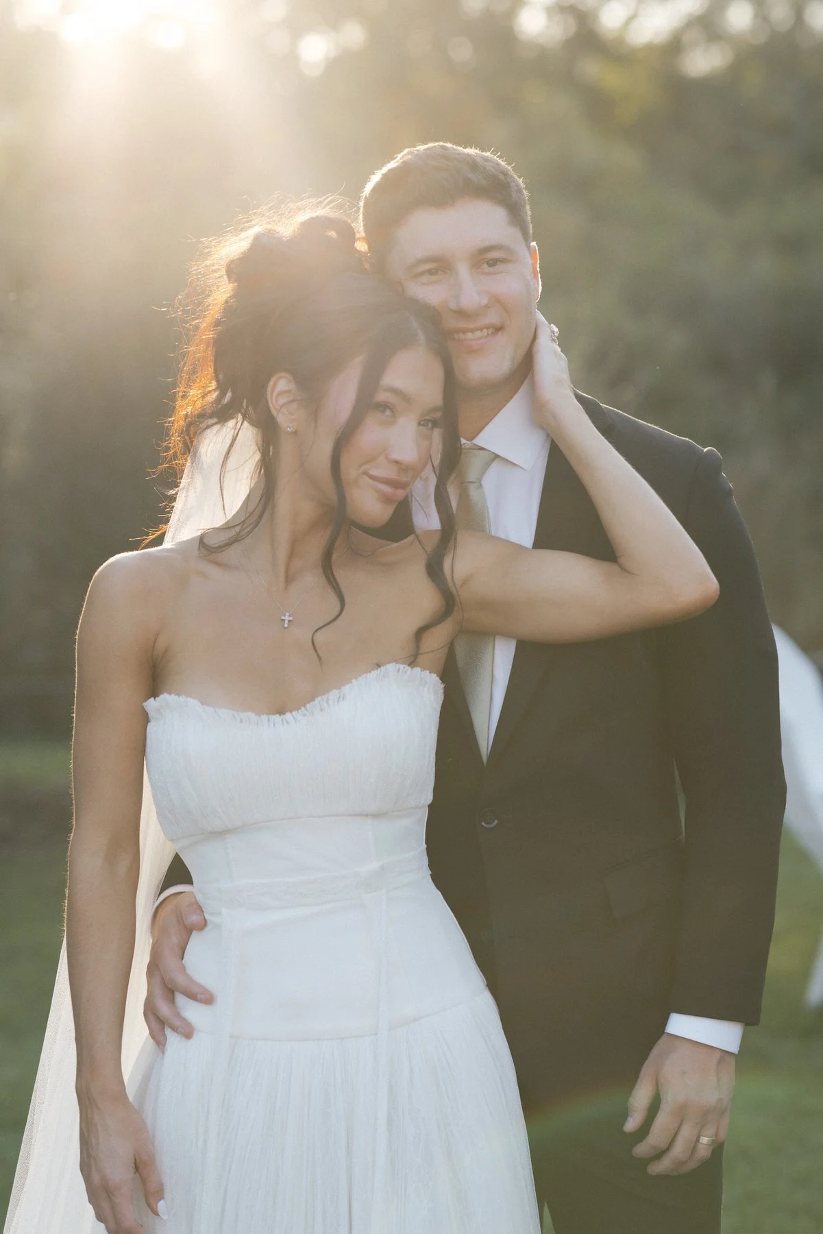 A bride and groom pose outdoors during sunset, the bride in a white strapless wedding dress and the groom in a black suit with a gold tie.