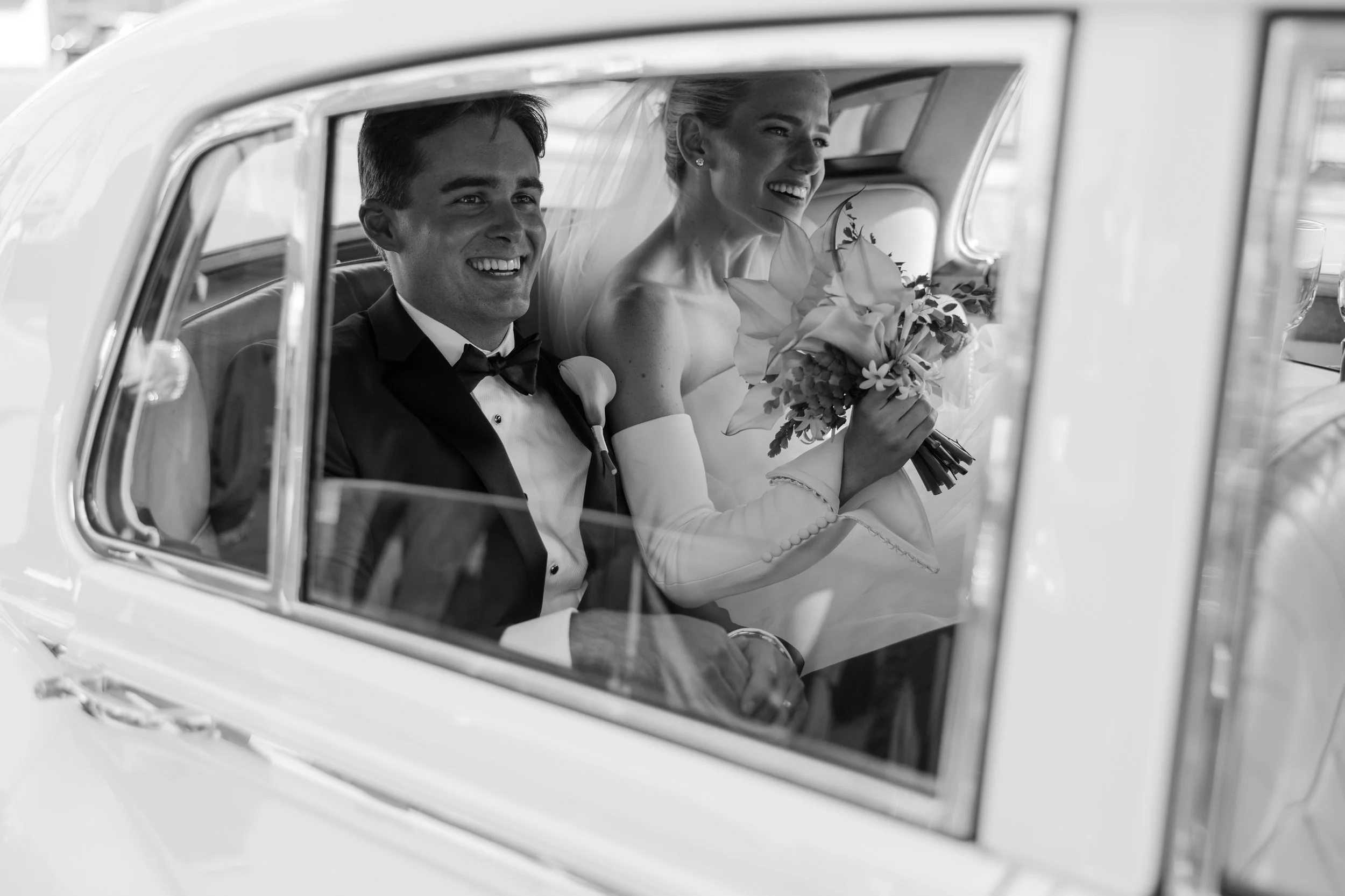 Black and white photo of a smiling bride and groom sitting inside a vintage car, with the bride holding a bouquet of flowers.