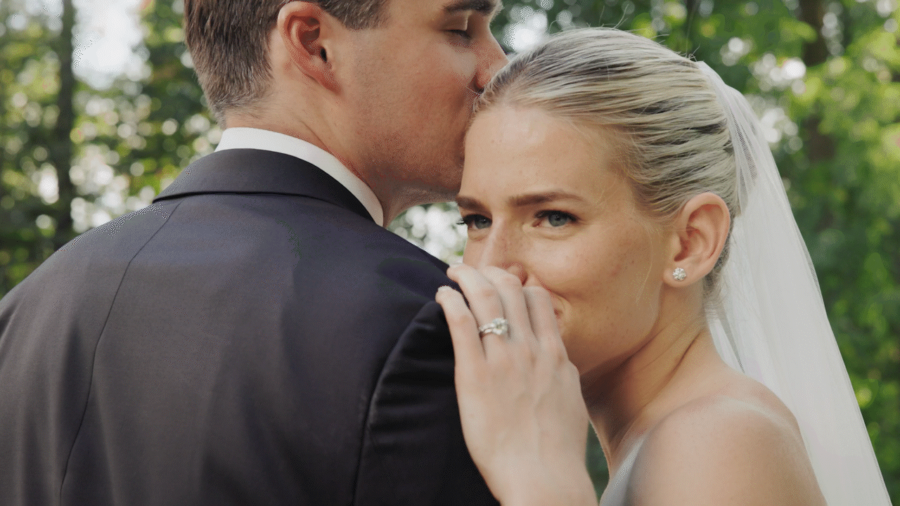 A bride and groom sharing a tender moment outdoors, with the groom kissing the bride on her forehead and the bride smiling softly, showing her wedding ring.