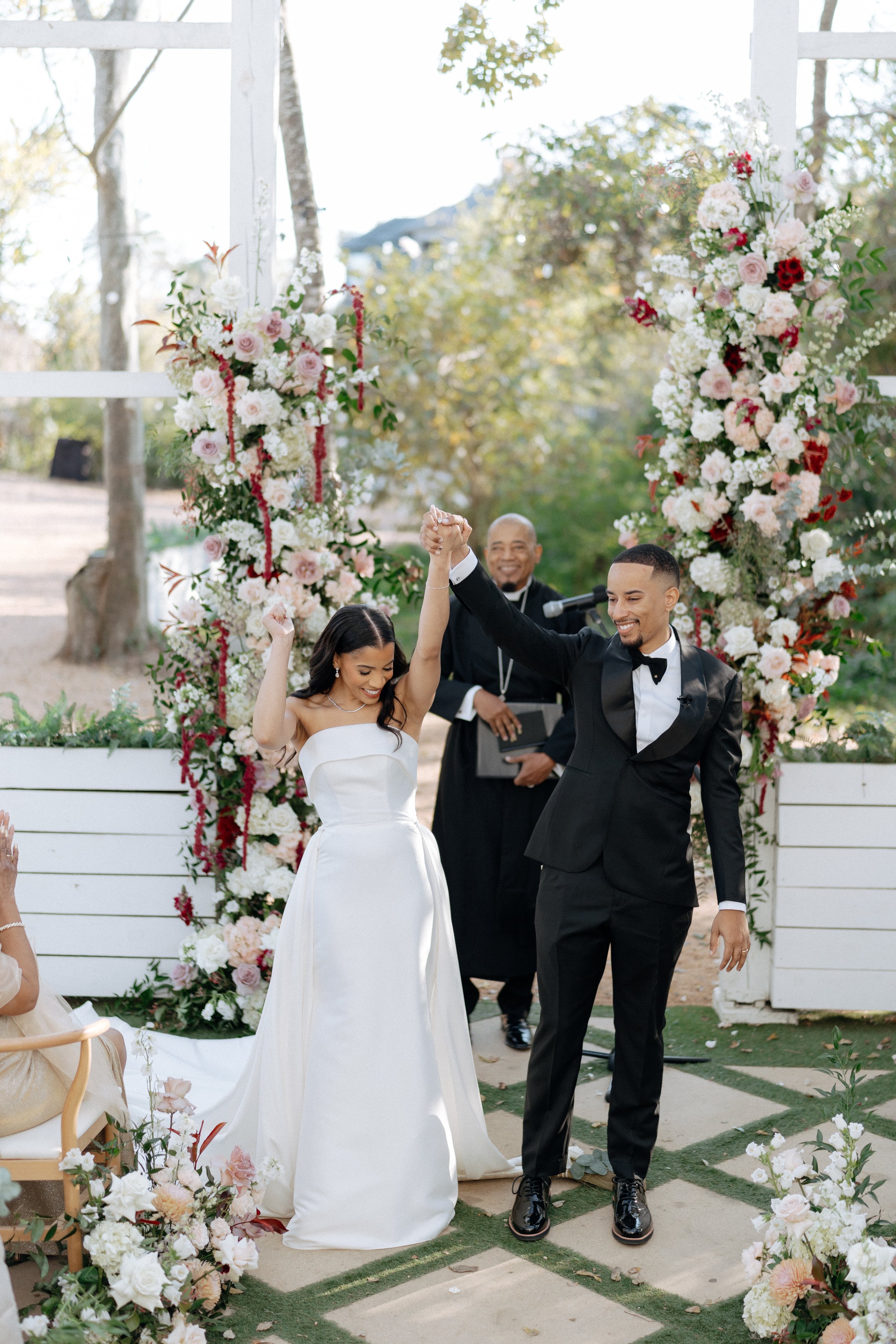 A newly married couple, the bride in a white wedding dress and the groom in a black tuxedo, celebrating at their outdoor wedding ceremony, holding hands and smiling in front of a floral arch with an officiant in the background.