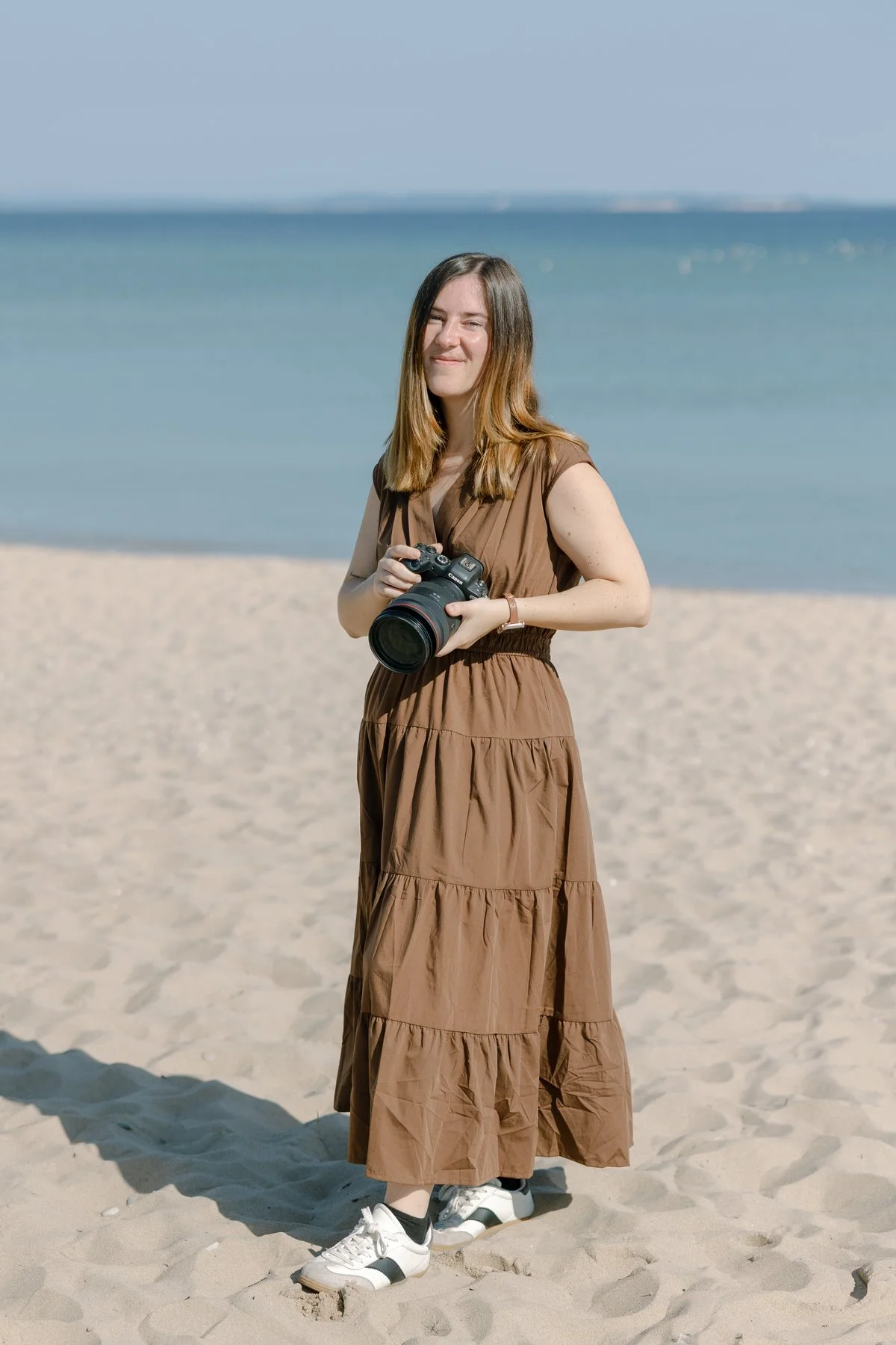 Jenna Horn in a brown dress holding a camera on a sandy beach with the ocean in the background.