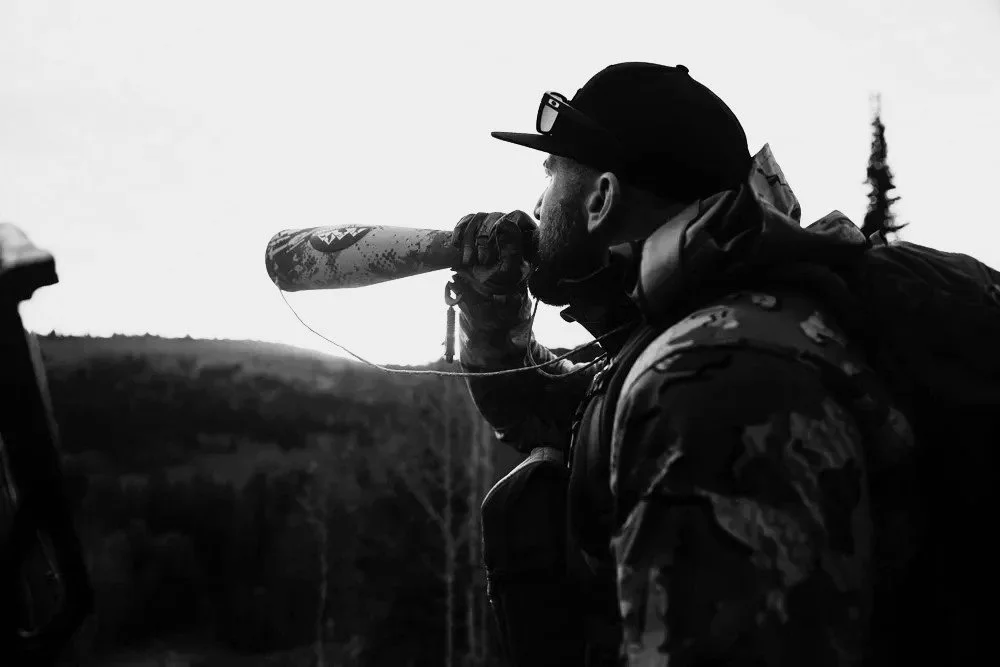 A man in camouflage clothing and a cap, outdoors, holding a rocket launcher to his shoulder, aiming it across a landscape of hills with trees, during sunset or sunrise.