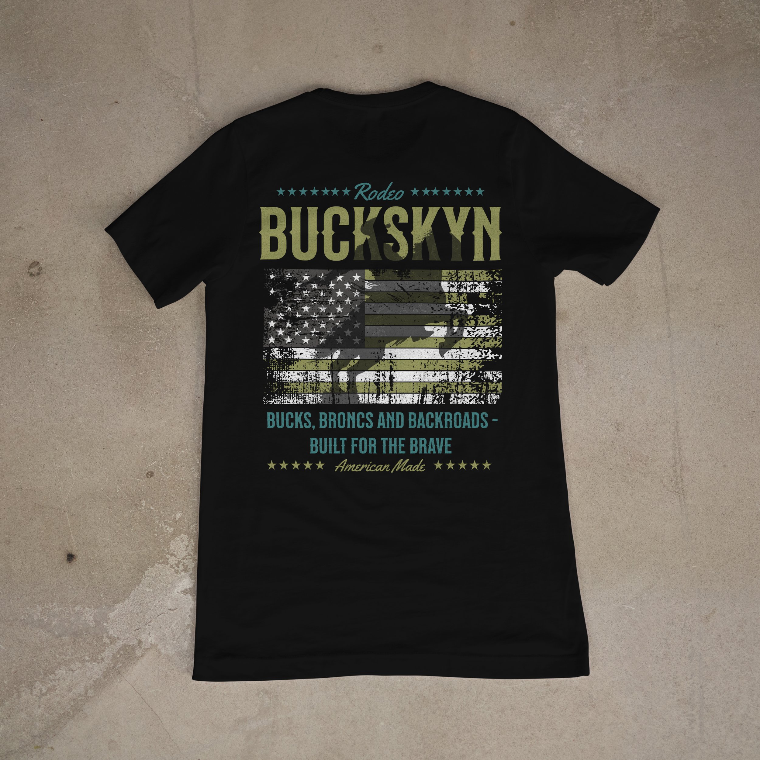 Black T-shirt with patriotic design featuring an American flag in camouflage pattern, with text 'Rodeo Buckskyn' and 'Bucks, Broncos and Backroads - Built for the Brave' in shades of blue and yellow.