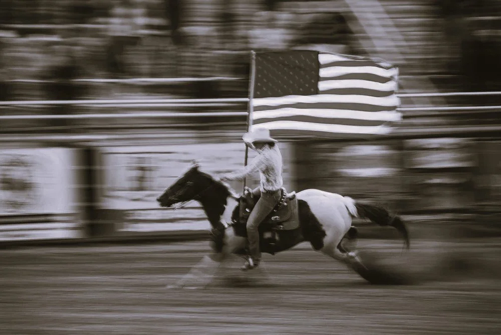 Person riding a galloping horse while holding an American flag in a blurred outdoor setting.