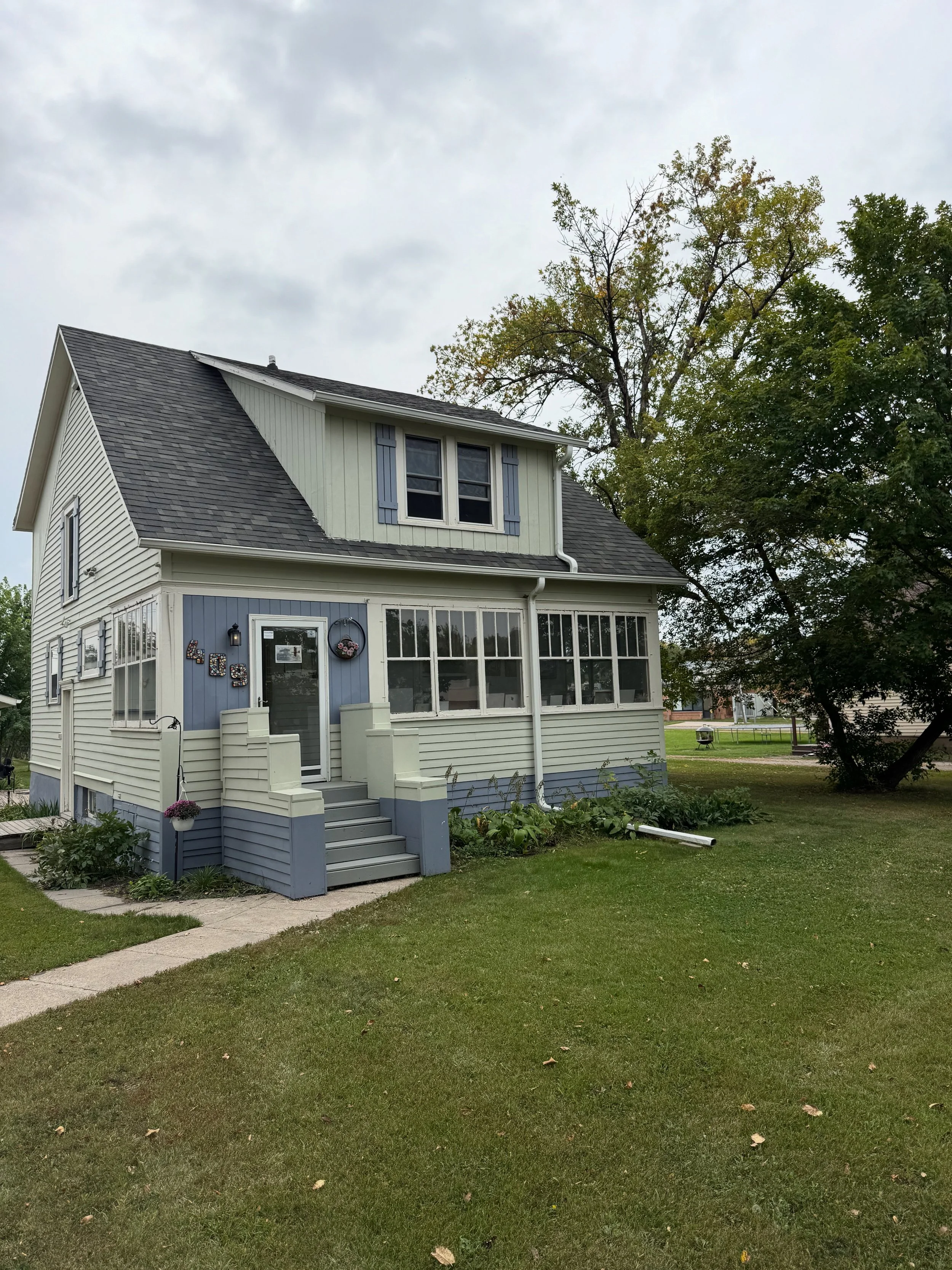 A two-story house with beige vertical siding, blue accents, and a front porch with stairs. The house is surrounded by green lawn and trees, with cloudy skies overhead.